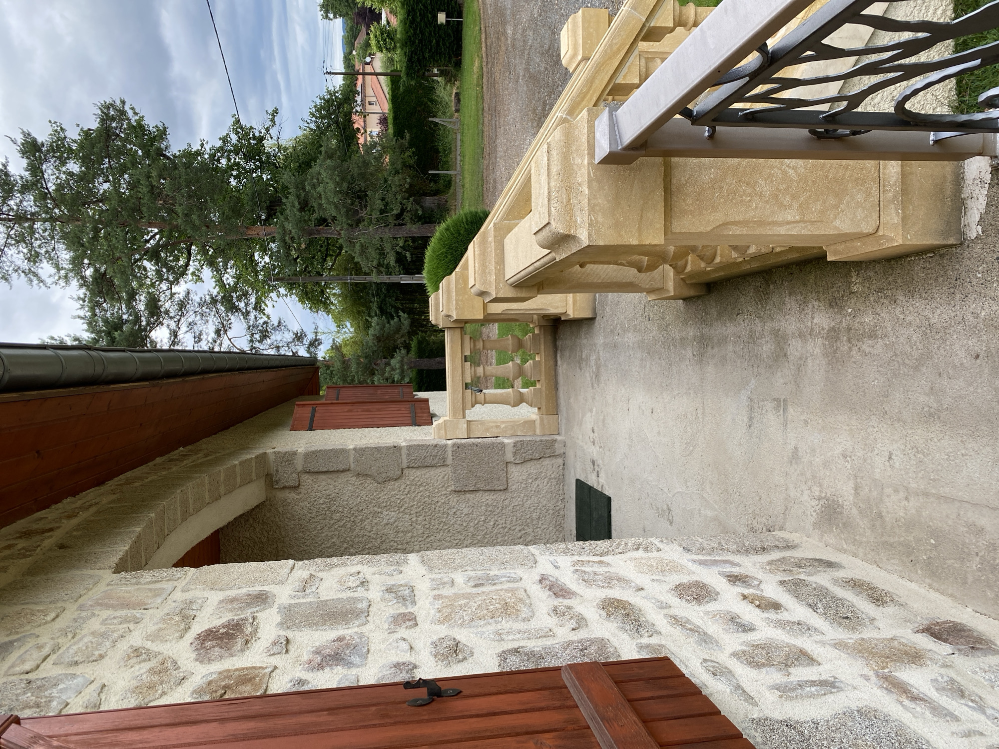 This media file presents an outdoor architectural scene in Courpière, France, under an overcast sky. The image primarily features the exterior of a building and an adjacent garden area.
On the left and in the foreground, part of a building is visible, characterized by walls constructed from varied, light-colored stone masonry. Dark reddish-brown wooden shutters are partially closed over windows, and a section of a brown-tiled roof or eaves is also present. An archway is subtly integrated into one of the building's walls.
To the right, a prominent and ornate balustrade crafted from light yellow/beige carved stone extends along a paved or gravel path. The balustrade is topped with a gray metal handrail featuring decorative, abstract scrollwork.
Beyond the immediate architectural elements, a vibrant green landscape unfolds. Several tall evergreen trees, likely pines, stand prominently, accompanied by various bushes and other smaller trees. In the distant background, residential structures with distinct orange-tiled roofs are nestled amidst the foliage, suggesting a rural or semi-rural setting. Utility poles and lines are faintly discernible against the cloudy sky, which displays streaks of white.
The scene is tranquil and devoid of human activity or visible text. The lighting indicates a bright, yet overcast, daytime, casting soft, diffused light across the landscape and structures.