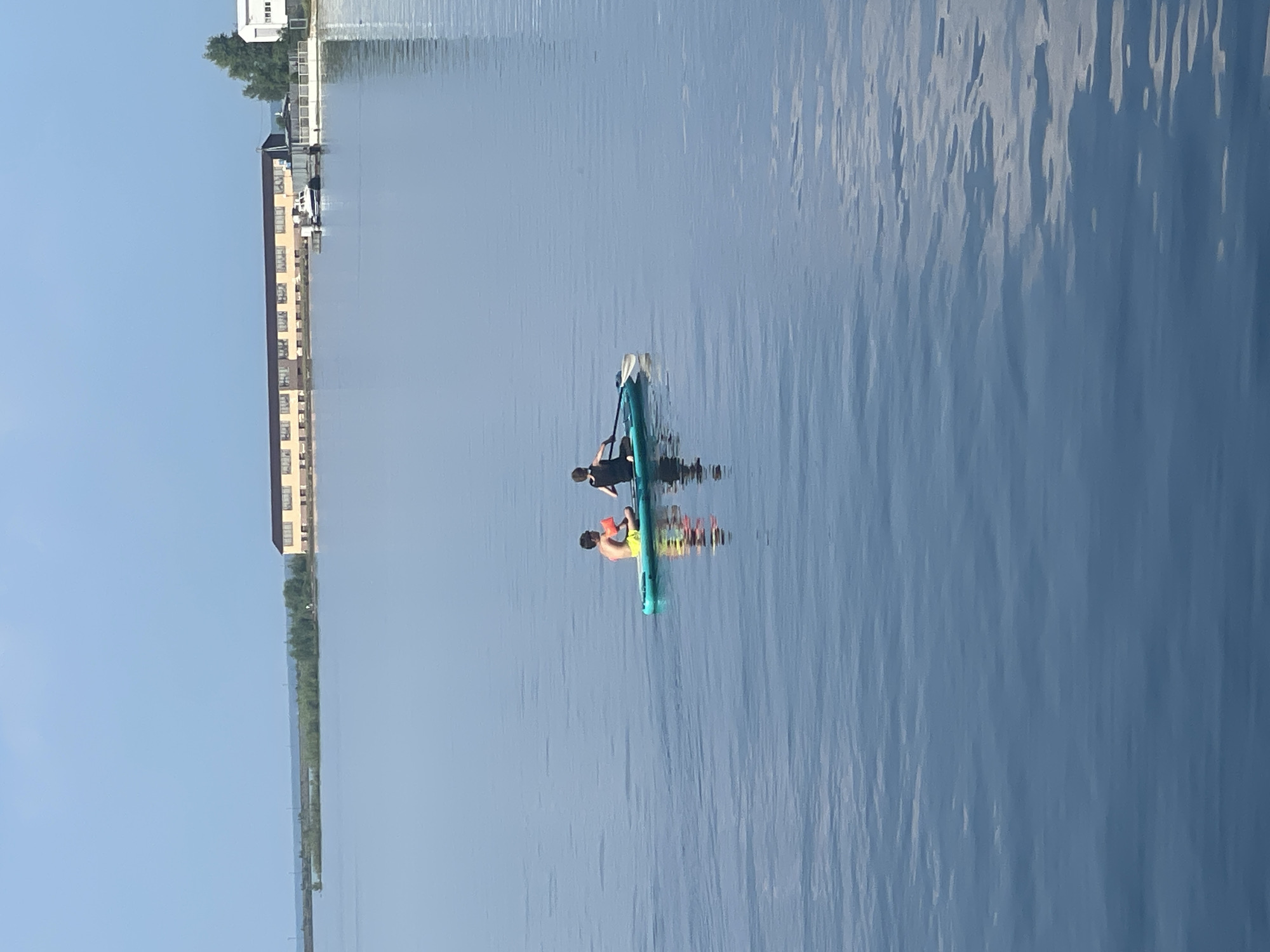 The image shows two individuals on a paddleboard in a body of water. One individual, wearing a yellow swimsuit and orange arm floaties, is sitting and holding a paddle. The other individual is kneeling and using a paddle to propel the board. The paddleboard is blue and white. In the background, there are several buildings with tan facades and dark roofs. A small white boat is docked near one of the buildings. The water is calm with subtle ripples. The sky is a clear blue. This scene is likely located on the Kama River in Chaikovsky, Russia, given the characteristic long, low buildings and the presence of the river.