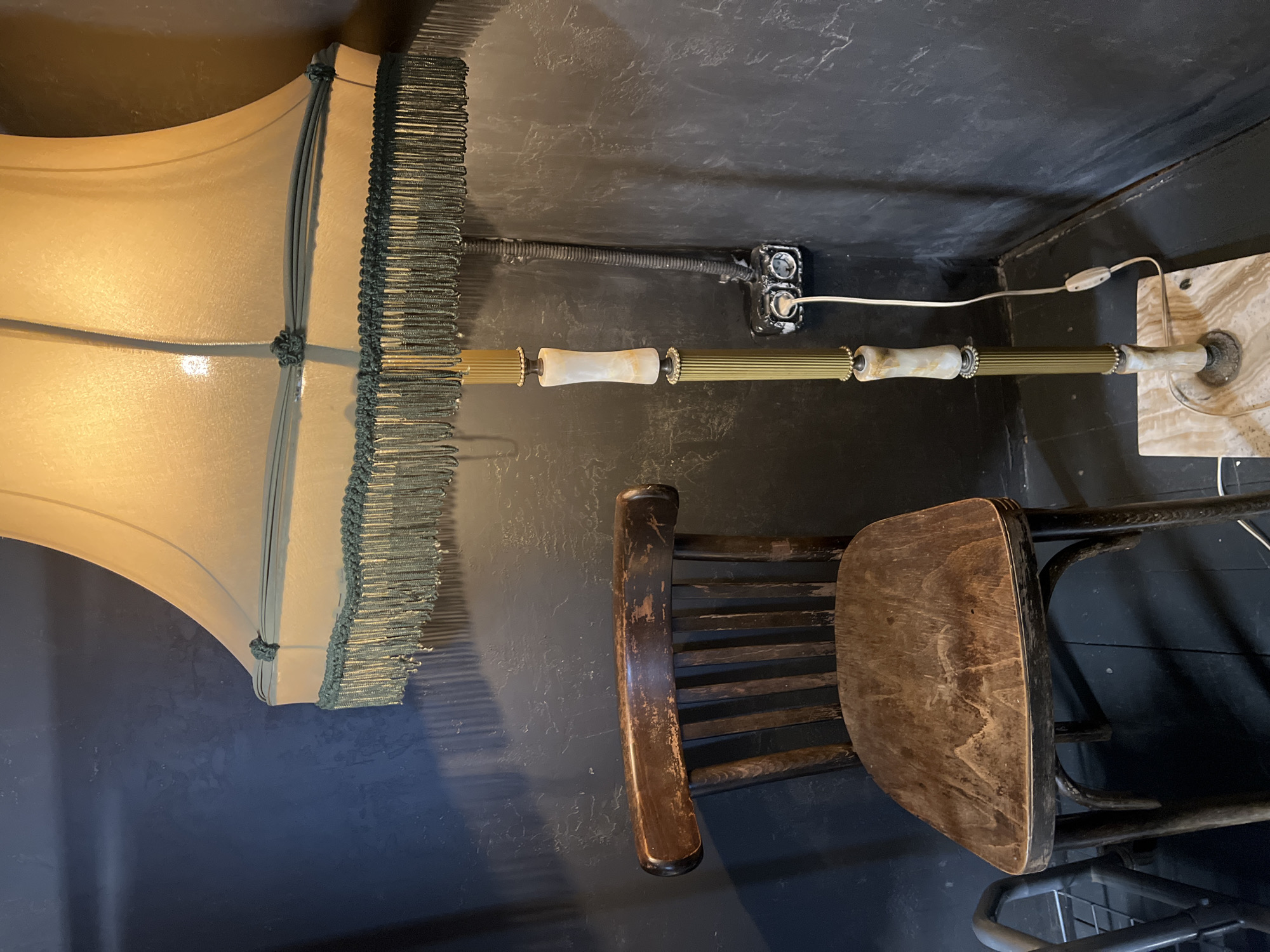 The image depicts a dimly lit interior corner. A standing lamp with a large, cream-colored shade adorned with green fringe is positioned in the corner. A worn, wooden chair sits nearby. The walls are a dark, textured gray. An electrical outlet with a black cover is visible on the wall, with the lamp's cord running along the wall toward the outlet. The lamp's base appears to be marble.
There is no information available in the image to indicate the location in the city of Moscow, Russia.