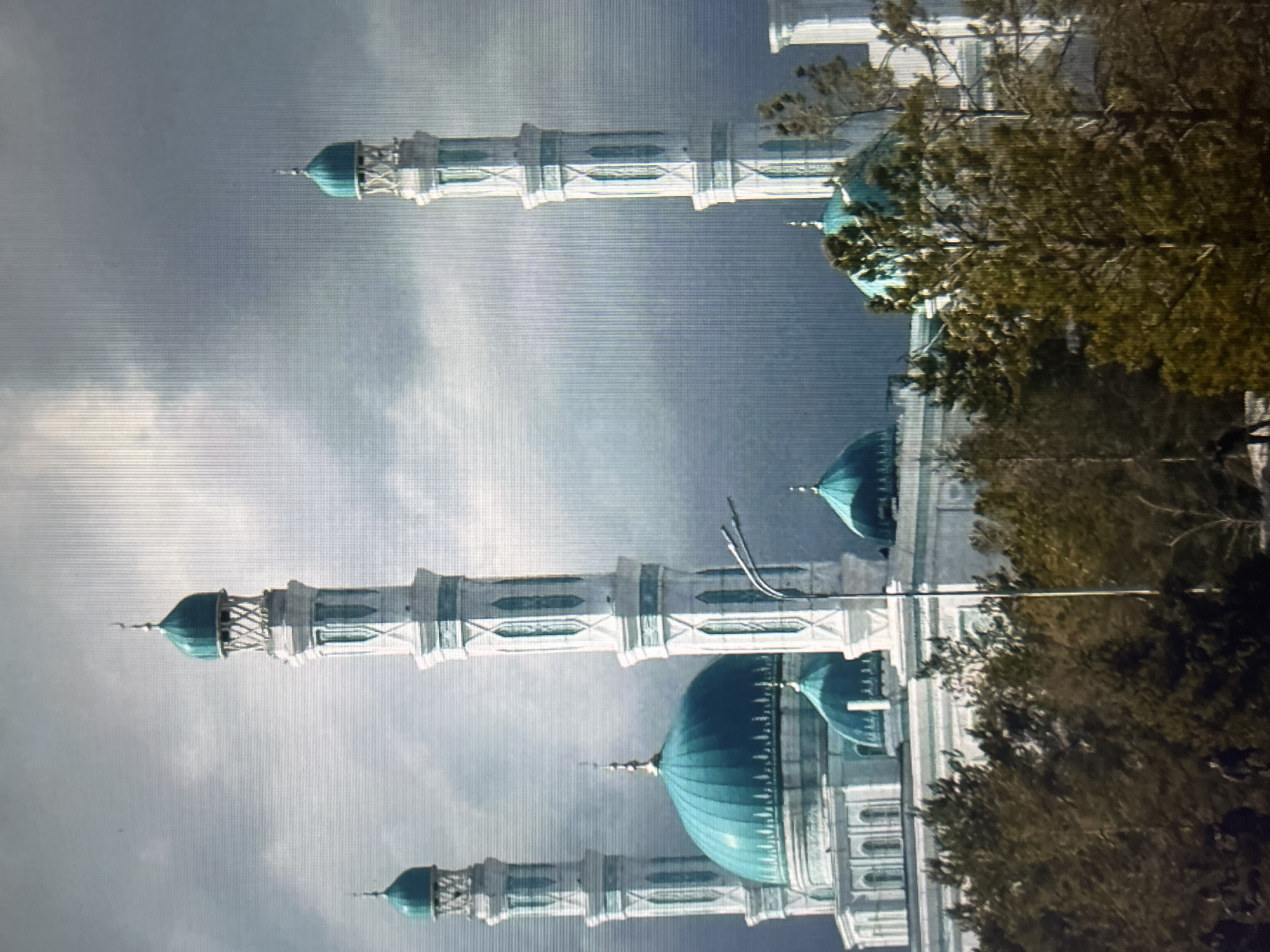 The image depicts a grand mosque with multiple minarets and domes, characterized by white facades with turquoise accents. The central part of the mosque features a large turquoise dome, flanked by smaller turquoise domes and four tall, slender minarets with turquoise conical tops. The architecture exhibits intricate detailing on the minarets and windows. In the foreground, a dense line of trees, predominantly coniferous, with some bare branches, partially obscures the lower section of the mosque. The sky is overcast with a mix of grey and white clouds, suggesting a possibly cool or dramatic weather condition. The image appears to be taken from a distance, capturing the impressive scale of the religious building. At the very bottom of the image, faint figures of people are visible amidst the trees, their actions unclear due to the distance and resolution.
