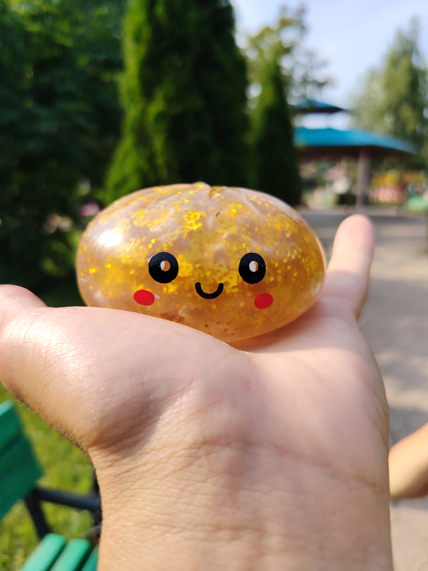 A close-up, eye-level shot shows a person's hand holding a small, yellow, translucent squishy toy filled with gold glitter. The toy has a simple, cartoonish face with two black eyes, a curved black smile, and two red circles on its cheeks. The background is softly blurred, revealing green trees, a pathway, and a gazebo with a blue roof. The lighting suggests it is daytime and outdoors, possibly in a park or recreational area in Chaikovsky, Russia.