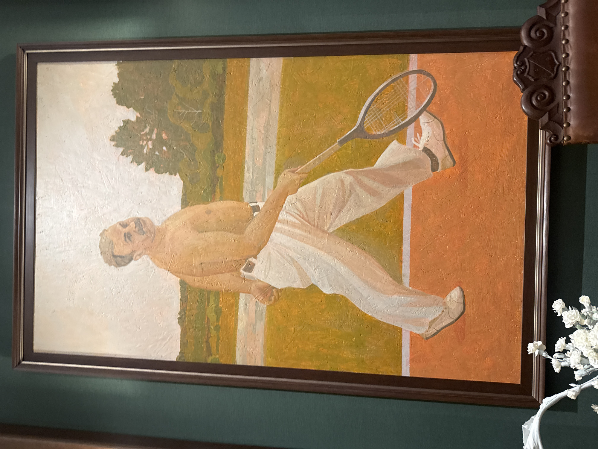 A painting depicts a man on a tennis court holding a racket. The man is bare-chested, wearing white pants and white tennis shoes. He has a mustache and short, light-colored hair. The tennis court is orange, with a white line marking its boundary. In the background, there is a field of yellow and green grass with trees. The painting is framed in dark brown wood and hangs on a dark green wall. The painting is located in the city of Zhukovka, Russia.