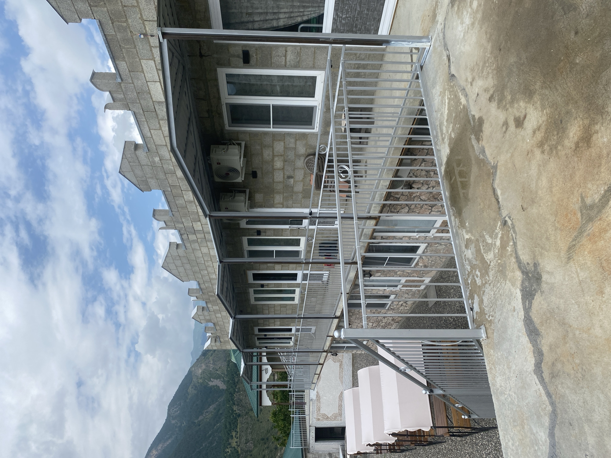 This is a medium-shot, eye-level, outdoor photograph taken in Chalda, Russia. The image features a multi-story building constructed with light-colored cinder blocks and stone. The building has a row of windows with white frames and a metal awning extending from the upper floor. The rooftop is adorned with decorative crenellations, resembling a castle's battlements. In the foreground, a concrete patio with cracks and water stains leads to a metal railing that encloses a walkway. Several white covered lounge chairs are visible on a lower level. In the background, a lush green mountain slopes upwards under a cloudy sky. The overall impression is one of a rustic or resort-like establishment nestled in a mountainous region. There are no people visible in the image.