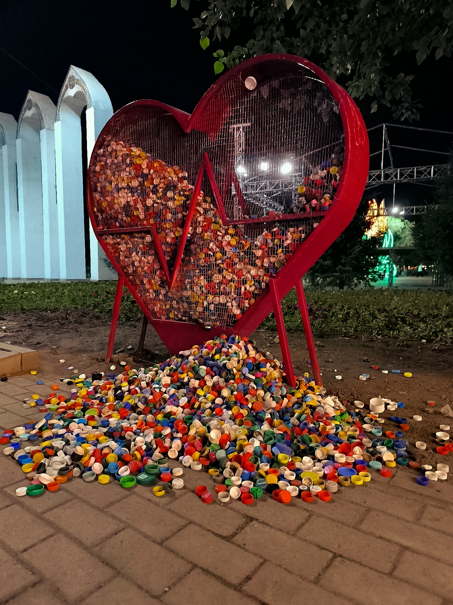 A large, red, heart-shaped sculpture filled with colorful plastic bottle caps stands in an outdoor area at night. A pile of loose bottle caps spills out from the bottom of the sculpture onto a paved walkway. The sculpture is situated in front of a white structure with arched openings and a dark, leafy tree overhead. In the background, there are lights and more trees, suggesting a park or public space. The image appears to be taken in Qarağandı city administration, Kazakhstan, though this is not explicitly depicted in the image itself.