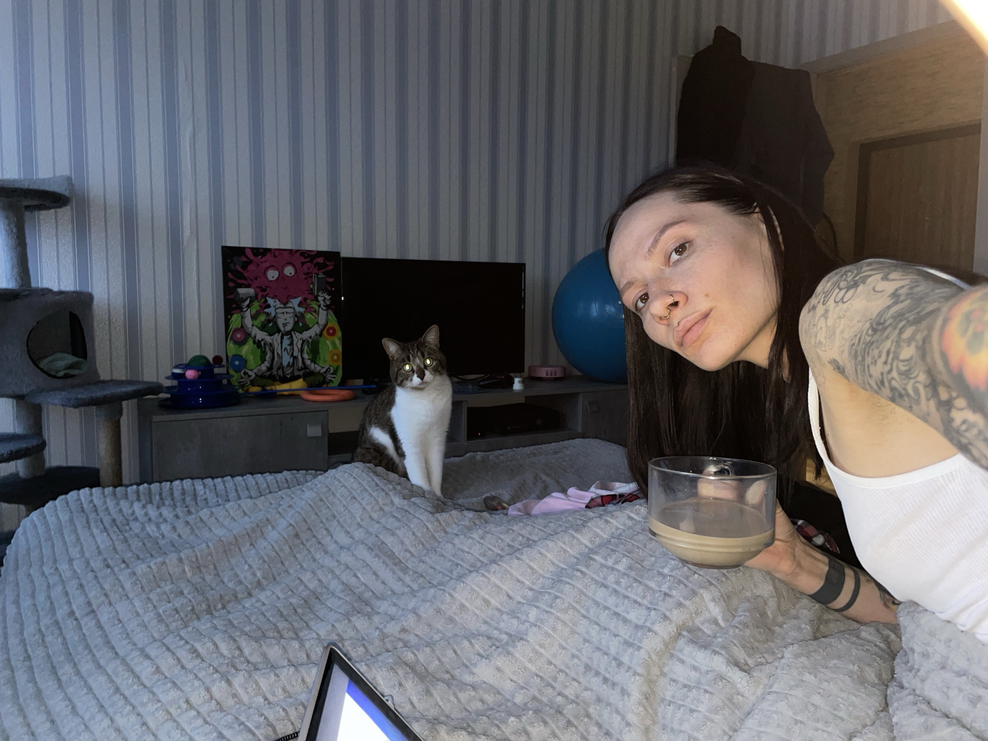 The image, oriented vertically, depicts an indoor scene specified as Izhevsk, Russia. A female individual is present in the lower right portion of the frame, positioned in a reclined or lying posture. She has long dark hair, visible tattoos on her left upper arm/shoulder area, and a black band tattoo on her right forearm. Her nose exhibits a septum piercing and an additional nostril ring. She is wearing a white tank top and gazes directly at the camera with a neutral expression. Her right hand holds a clear glass containing a light brown liquid.
To the left of the female, a brown and white tabby cat is partially visible, positioned behind a light grey, textured blanket that covers a bed. The cat's eyes reflect light, appearing bright yellow-green. A digital tablet with an illuminated screen is partially visible on the bed surface in the upper left.
The background wall features a light-colored striped wallpaper pattern. In the upper right corner, a multi-level grey cat tree is visible. Below it, a black flat-screen television is mounted on the wall. Above the television, a colorful poster depicting stylized, cartoon-like figures in predominantly pink and green hues is displayed. A partially deflated blue exercise ball rests on the floor in the lower-middle background. Other indistinct items, resembling clothing, are visible near the base of the bed.
