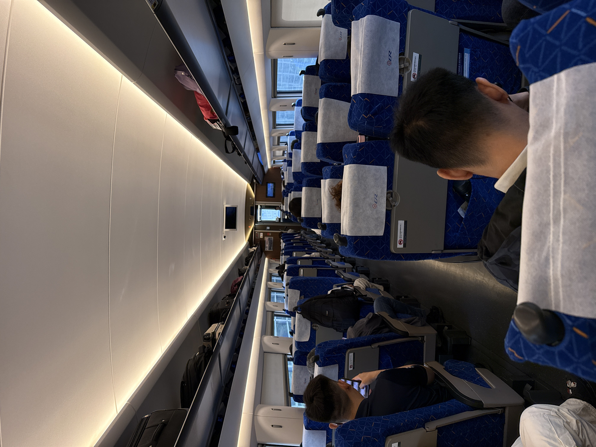 The interior of a train car is depicted, showing rows of blue and white patterned seats filled with passengers. Luggage is stored in overhead compartments. The lighting is bright, particularly along the ceiling. One passenger is using a phone, while others are seated. The location is unknown.