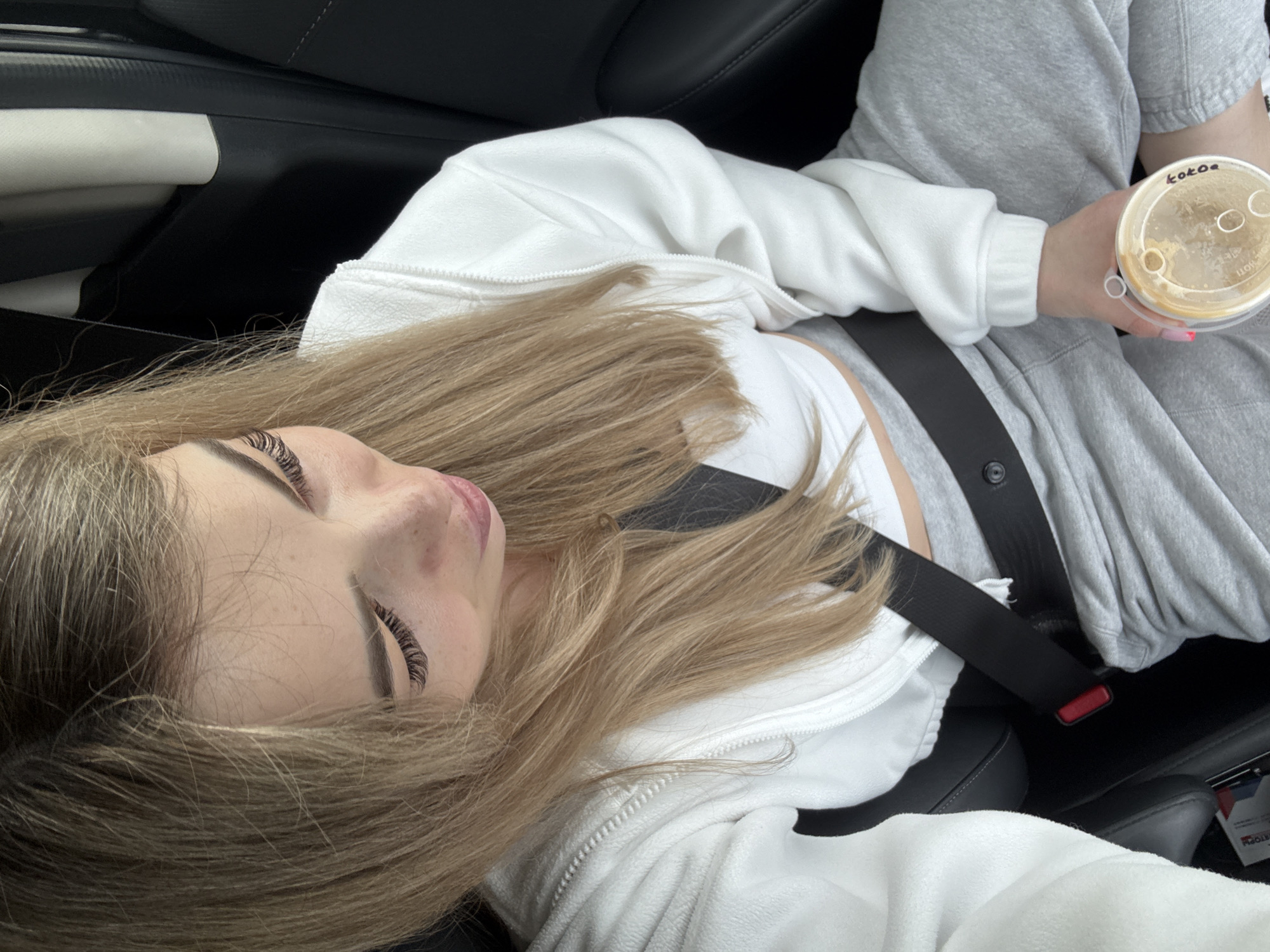 A woman is sitting in a car, wearing a white hoodie, grey sweatpants, and a white crop top, and is buckled in with a black seatbelt. She has long blonde hair and appears to be holding a coffee cup with "kokoa" written on the lid. The location is unspecified.