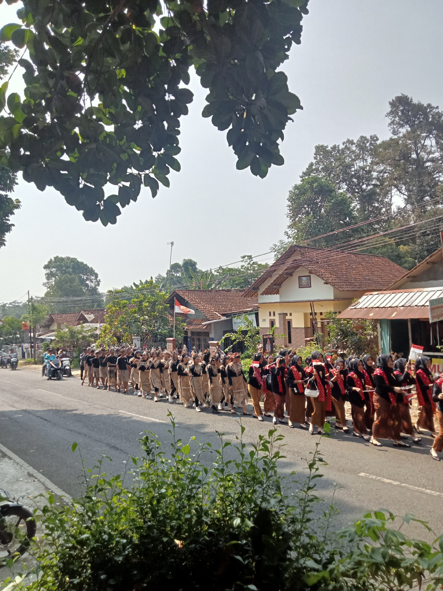 A group of people are marching in a parade on a street in Dororejo, Indonesia. The parade participants are wearing traditional clothing. Some are wearing brown skirts and black tops with red accents, while others are wearing black t-shirts and brown knee-length skirts. The group is walking on the right side of the road, which is paved. There are houses and trees lining the street. A few motorcycles are also visible on the road. The sky is bright and sunny.