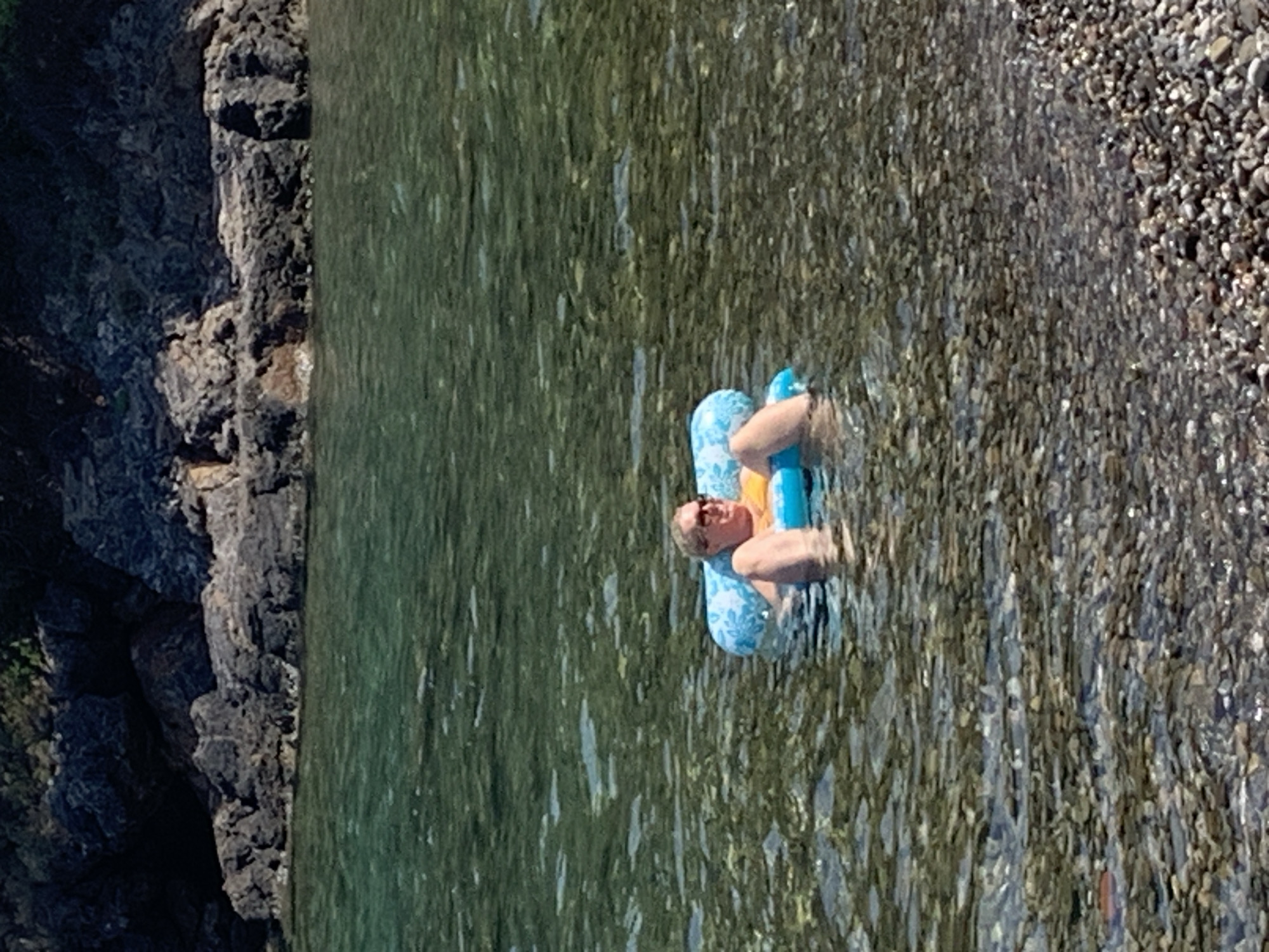 A woman is floating on an inflatable raft in the clear, shallow waters of Pozzarello, Italy. The raft is blue and white with a floral pattern. The woman is wearing sunglasses and an orange swimsuit. She is lying on her back, with her legs extended. The water is calm and transparent, revealing the rocky seabed. In the background, there are rocky cliffs and lush green vegetation. The sun is shining, casting a warm glow on the scene.