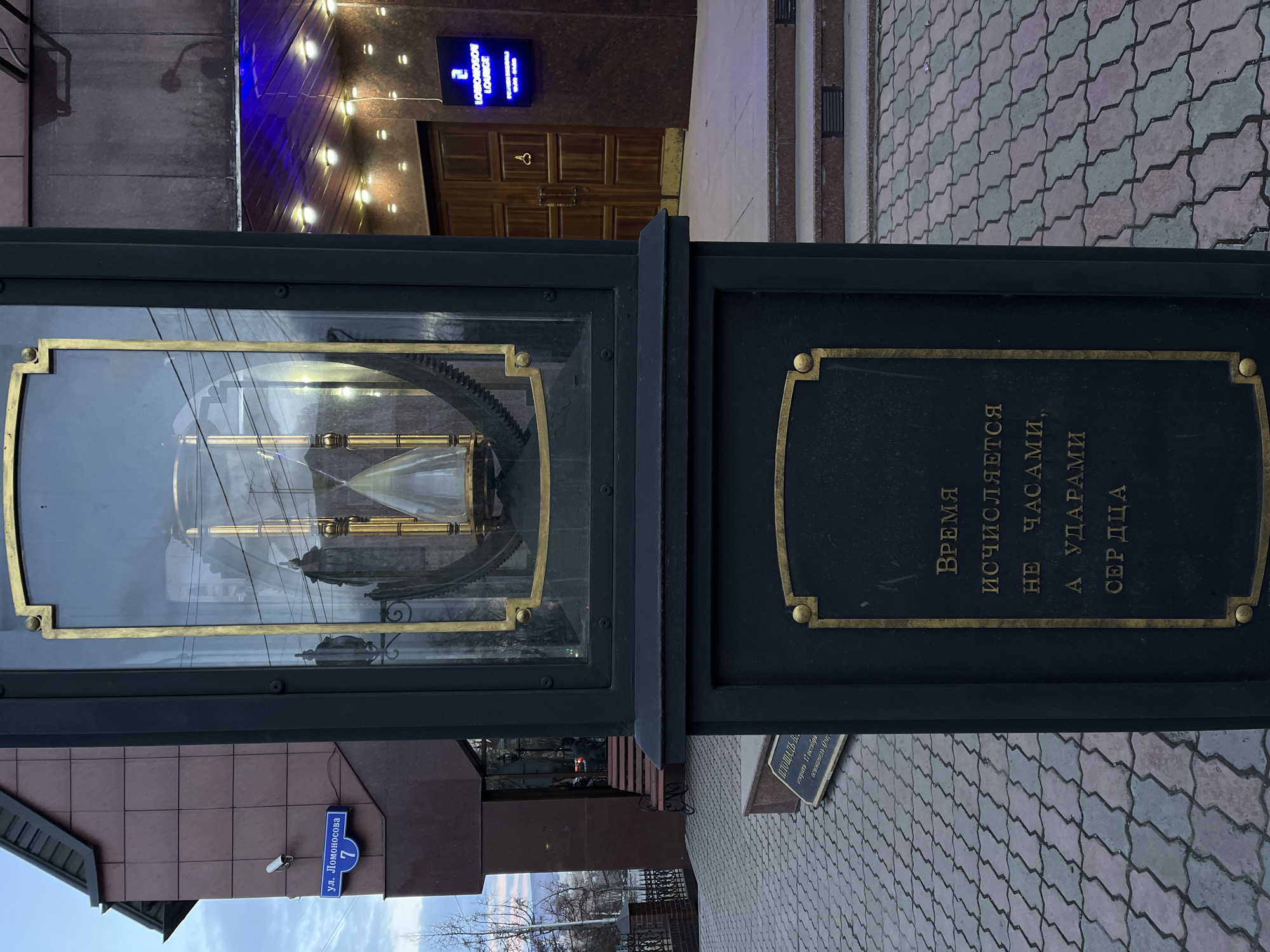 This vertically oriented image captures a detailed street scene in Krasnoyarsk, Russia, featuring a unique outdoor monument or street clock. The central object is a tall, dark-colored structure with golden trim, housing visible golden clockwork mechanisms behind glass, though no clock face is present.
A prominent plaque on the side of the monument bears a philosophical inscription in Russian: "ВРЕМЯ ИСЧИСЛЯЕТСЯ НЕ ЧАСАМИ, А УДАРАМИ СЕРДЦА" (Time is not measured by clocks, but by heartbeats). Closer to the ground, a second plaque is partially visible, stating "ПЛОЩАДЬ ВА открыта 17 октября основатель Олег" (VA Square opened October 17, founder Oleg).
To the left of the monument, a blue street sign clearly indicates "ул. Ломоносова 7" (Lomonosov Street 7), providing a precise location cue. In the background, a building with a brown-tiled facade hosts a modern, glowing blue sign that reads "LOMONOSOV LOUNGE" and the number "2". The area around the monument is paved with patterned interlocking stones in shades of grey and muted purple. The sky is bright, suggesting clear daytime weather. No individuals are visible in the scene.