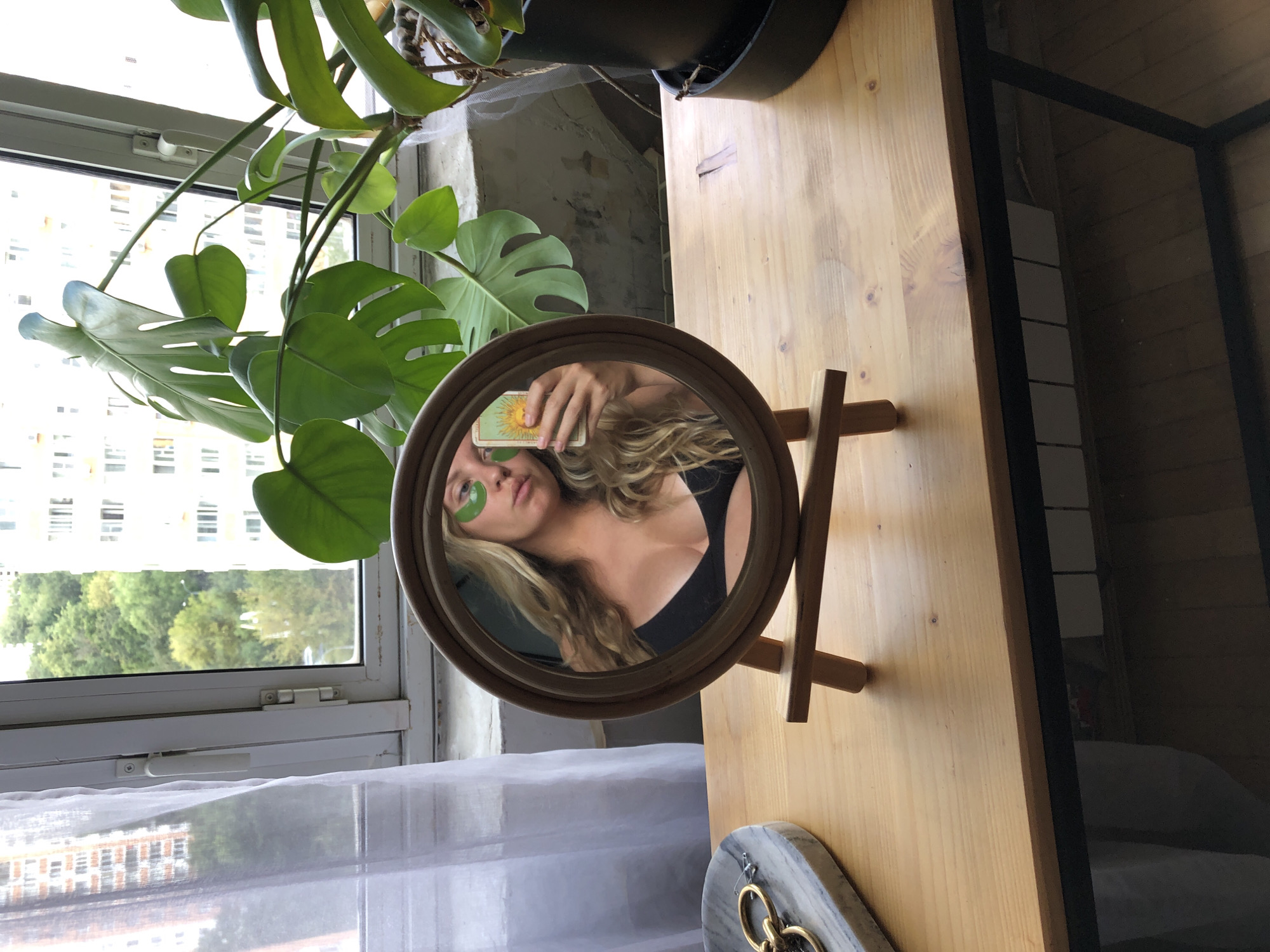 A blonde woman is pictured taking a selfie in a circular mirror. She is wearing a black bikini top and green eye patches. She is holding up a tarot card featuring the sun. The mirror is on a wooden stand, placed on a wooden table. To the left of the mirror is a marble dish with jewelry. Behind the mirror, a large monstera plant is visible. The scene is set against a window with a view of buildings and trees, indicative of an urban environment. The woman's reflection suggests she is indoors, by a window.