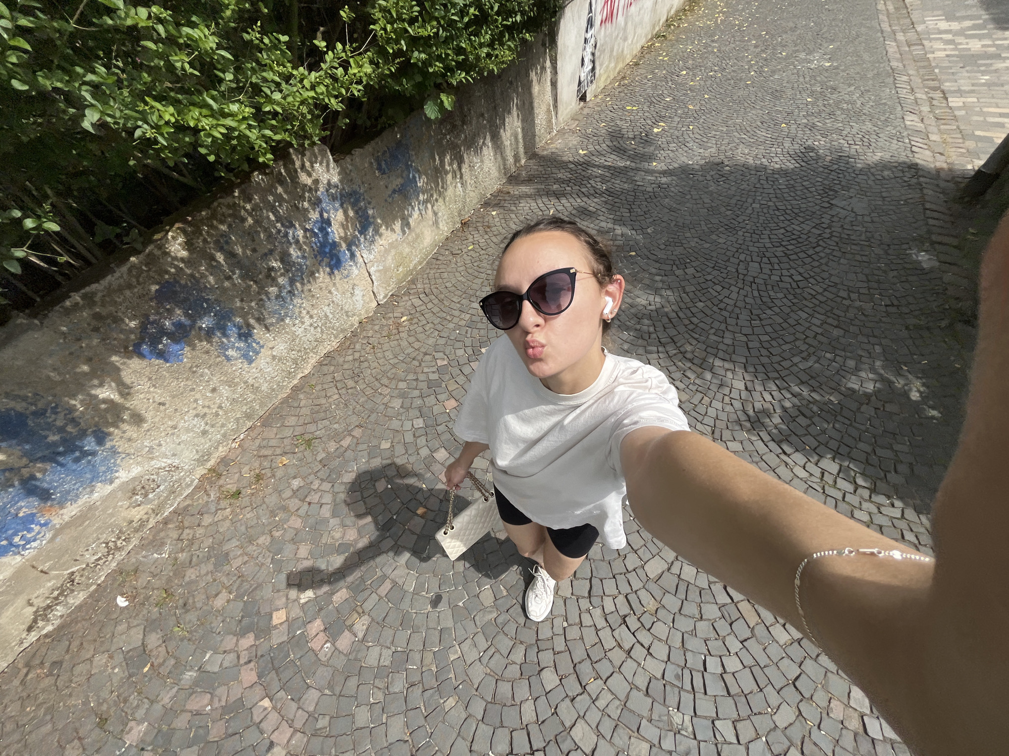 A young woman takes a selfie on a cobblestone street in Saarbrücken, Germany. She wears a white T-shirt, black shorts, white sneakers, and sunglasses, and has a white purse with a gold chain. She is making a kissing face. The background includes a wall with blue paint and green foliage. The street is paved with cobblestones.