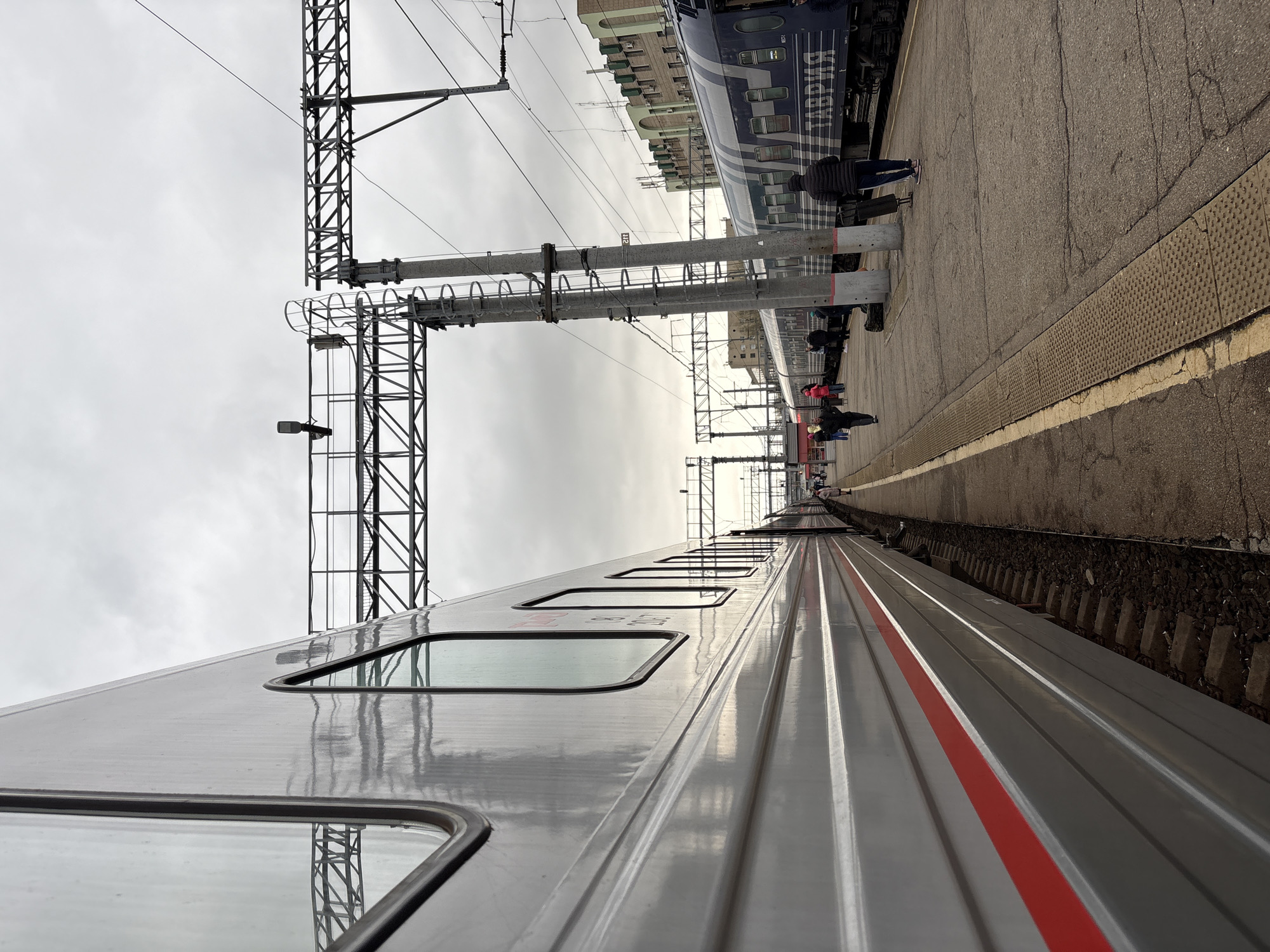 The image shows a train station in Saratov, Russia. The scene is overcast with a gray sky. Two trains are visible. One is a light-colored train with windows and a red stripe running horizontally along its side. The other train, located on a parallel track, is blue with white writing. Several people stand on the platform between the trains, some with luggage. Metal support structures for the overhead power lines are visible above the trains. The platform has a textured surface near the train and a concrete barrier along the edge of the platform.