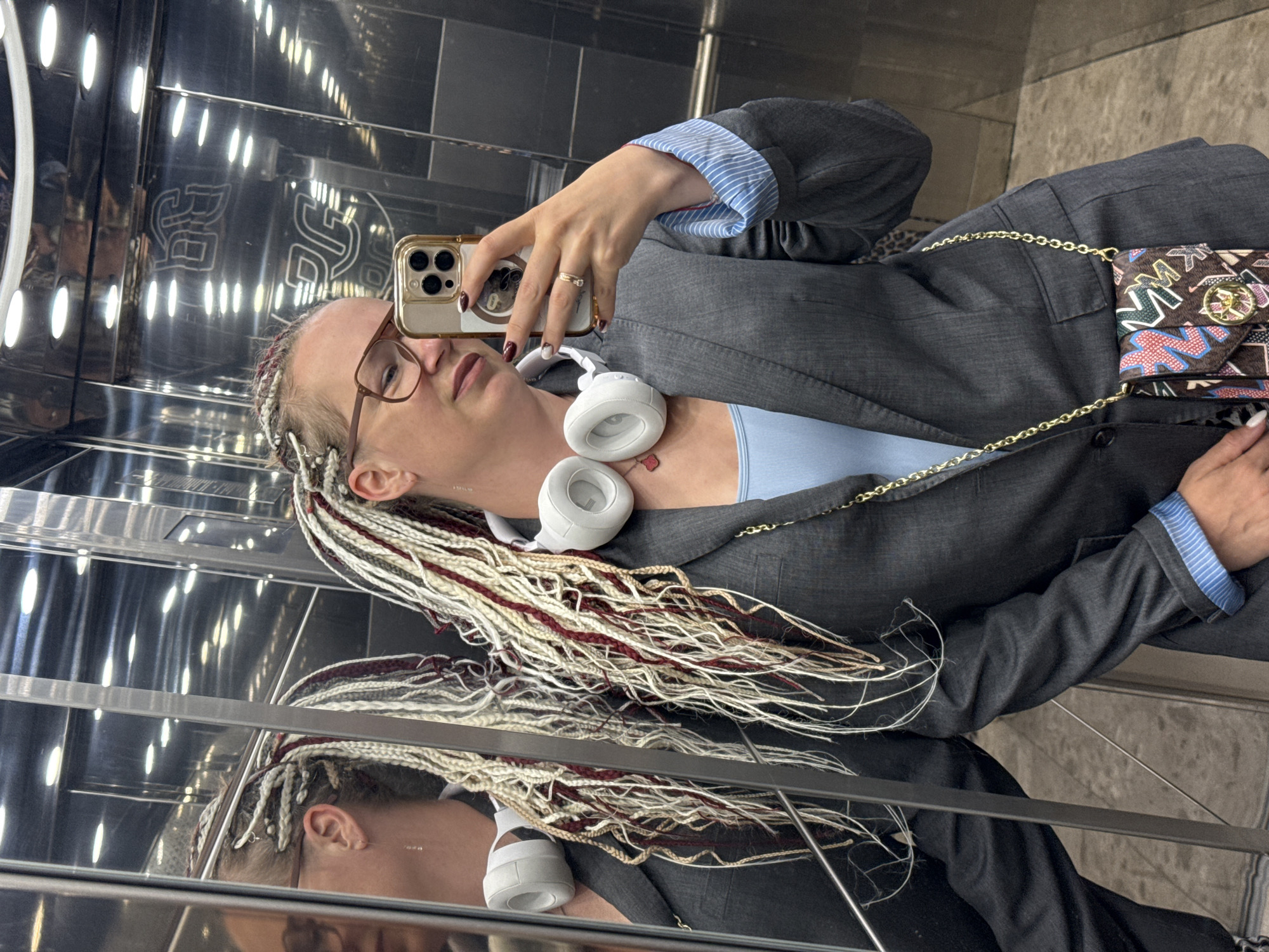 This is a vertically oriented, eye-level selfie taken by a woman in what appears to be an elevator. She is wearing a dark grey blazer over a light blue top, and a blue and white striped shirt is visible at her wrist. White headphones with a gold chain are draped around her neck. She has blonde braided hair with streaks of burgundy and is wearing glasses. A small, patterned purse with a gold chain is slung across her body. The elevator's walls are highly reflective, showing numerous lights and what appears to be a decorative logo or pattern. Her reflection is visible throughout the image. She is holding a gold-cased smartphone, capturing the selfie. The lighting in the elevator is bright and artificial, reflecting off the metallic surfaces. The overall mood seems casual and self-assured. There is no discernible time of day or weather information.