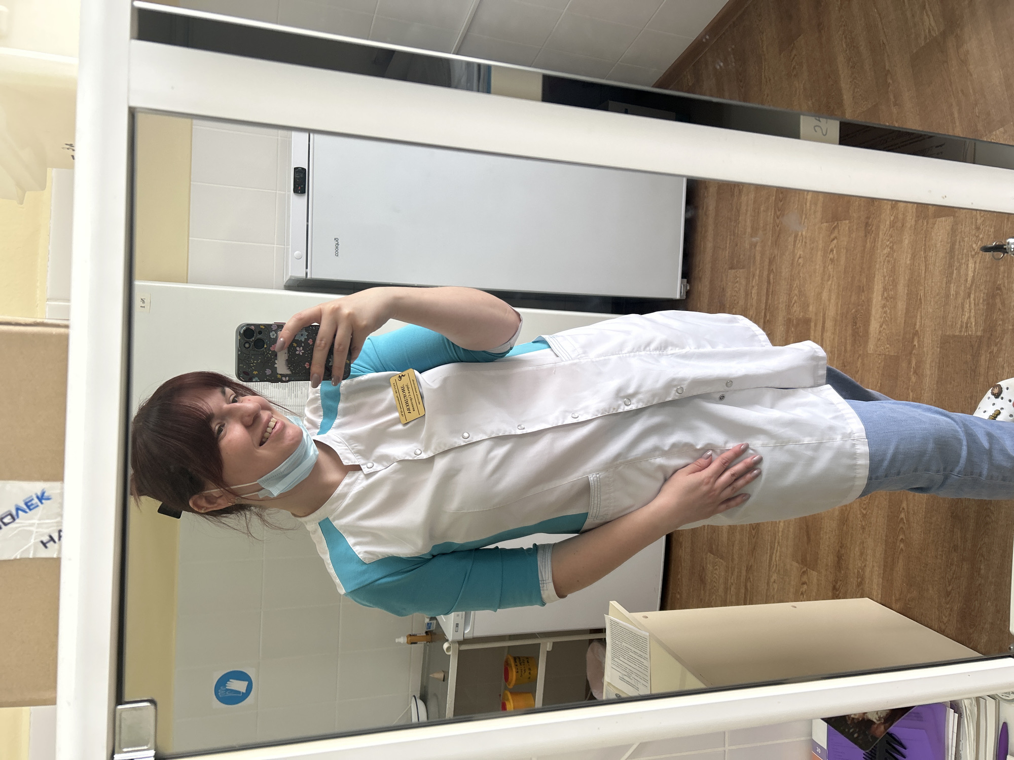 A young woman is taking a selfie in a mirror. She is wearing a light blue long-sleeved shirt with a white lab coat over it, jeans, and a blue surgical mask is partly covering her face. The photo is taken indoors in what appears to be a medical office or clinic setting. A refrigerator and a cabinet are visible in the background. A box is located near the mirror. I cannot determine the location in the city of Фоки, Russia.