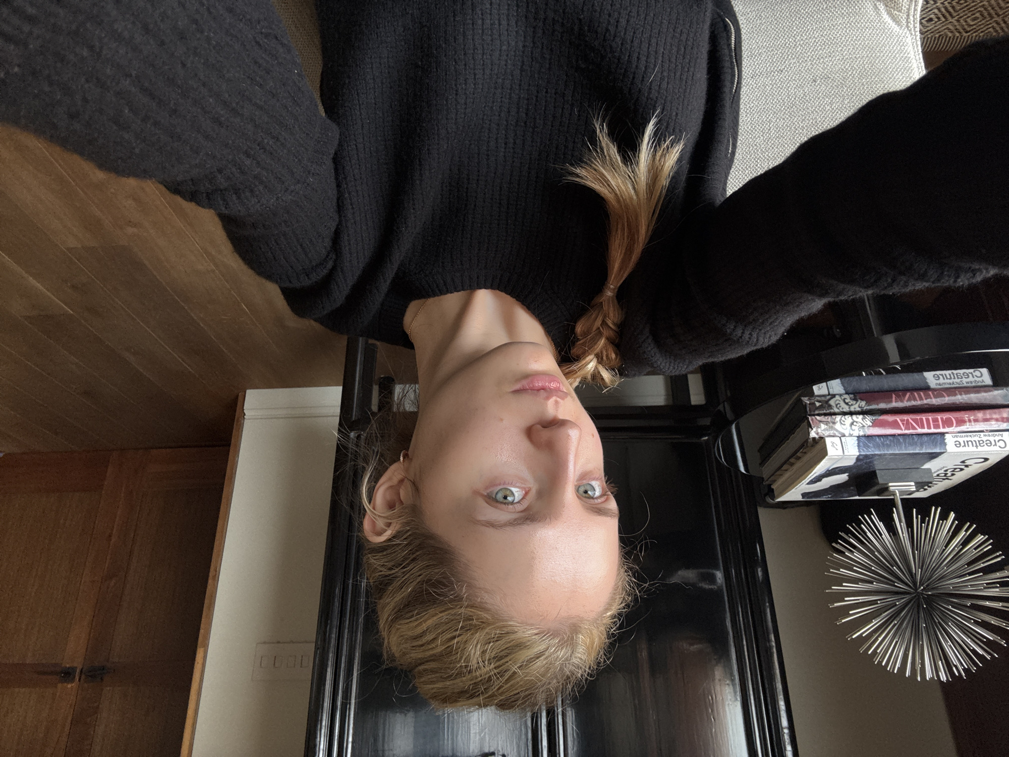 The image is an upside-down selfie of a woman with blonde hair, partially braided. She is wearing a black sweater. Behind her, there is a wooden floor, a cream-colored wall with an electrical outlet, and a dark-colored table with books and a silver decorative object on it. The spine of one of the books reads "Creature" by Andrew Zuckerman, and another reads "CHINA". The location in the city of Mexico City, Mexico cannot be determined from the image.