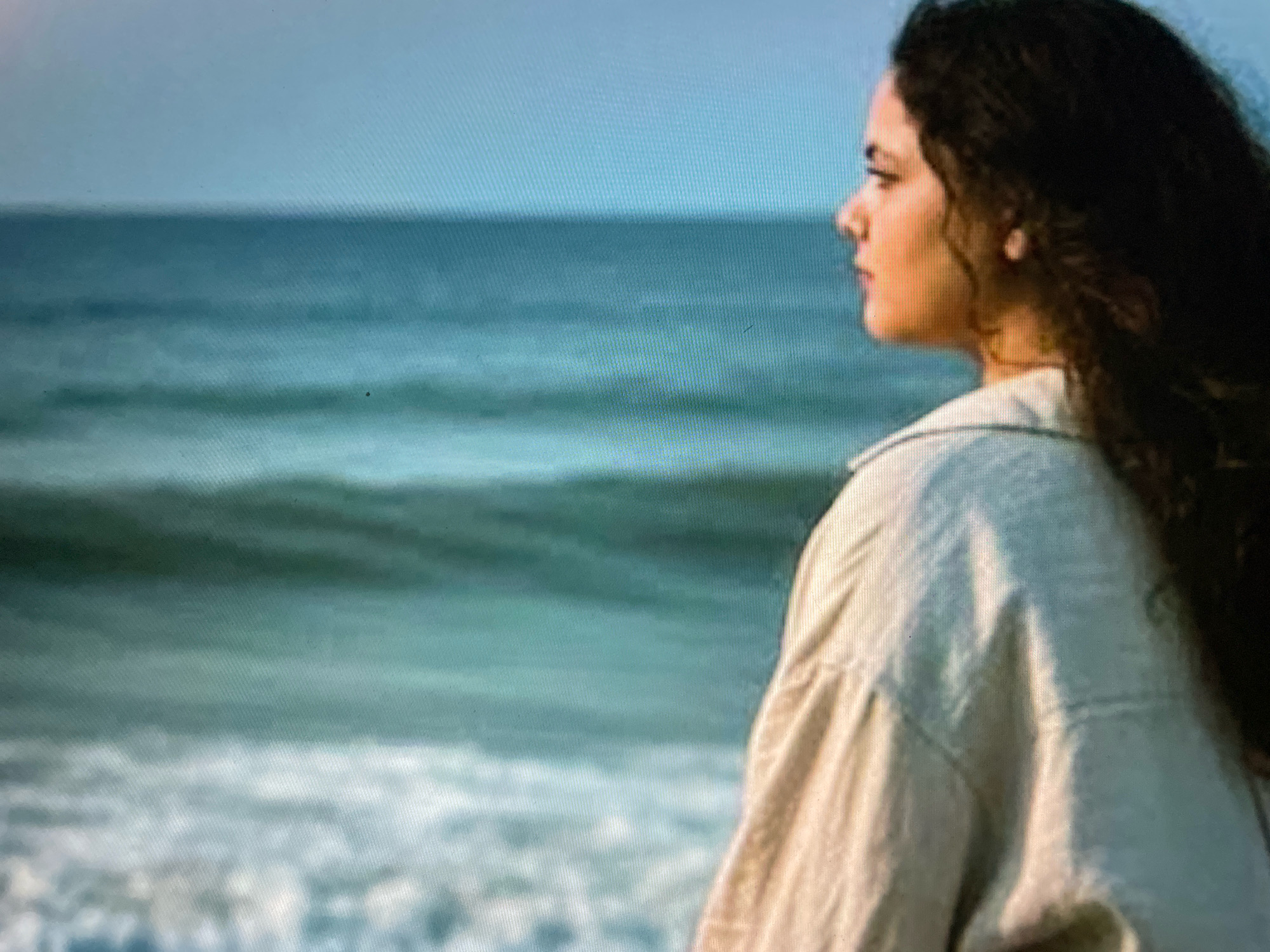 A young woman with dark, curly hair is shown in profile, looking out at the ocean. She is wearing a light-colored, collared shirt with puffy sleeves. The water is a greenish-blue, with gentle waves rolling towards the shore. The sky above is a clear, pale blue. The scene appears to be a peaceful coastal setting. There is no indication of the city of Gankar, India.