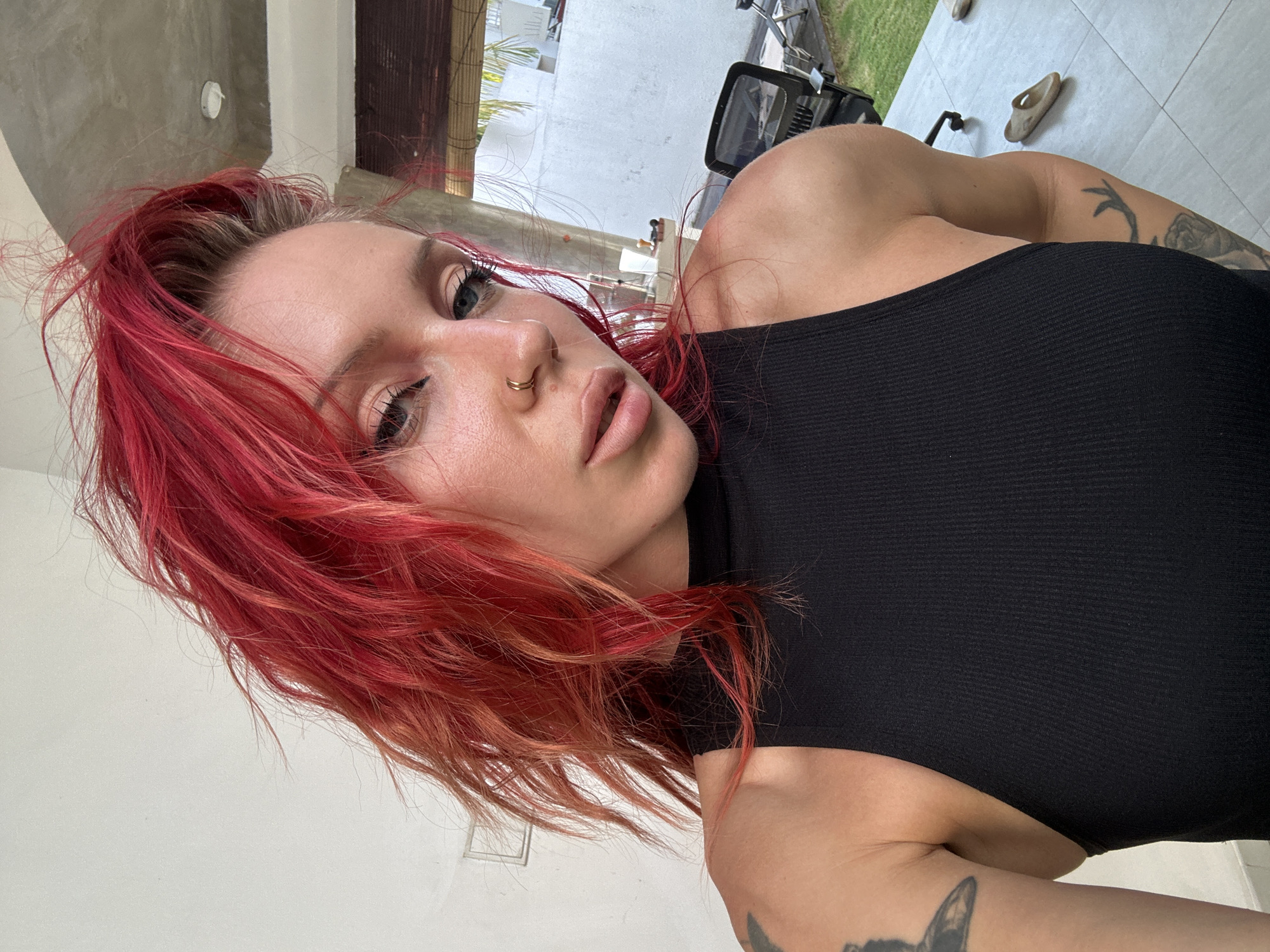 A woman with bright red, messy hair and blue eyes is captured in a close-up, selfie-style shot. She is wearing a black, ribbed tank top and has a gold hoop nose ring. Her left shoulder and arm are visible, revealing tattoos. The background suggests an outdoor setting, possibly a patio or yard, with a white wall, some greenery, and outdoor furniture visible. The scene appears to be in North Kuta, Indonesia.