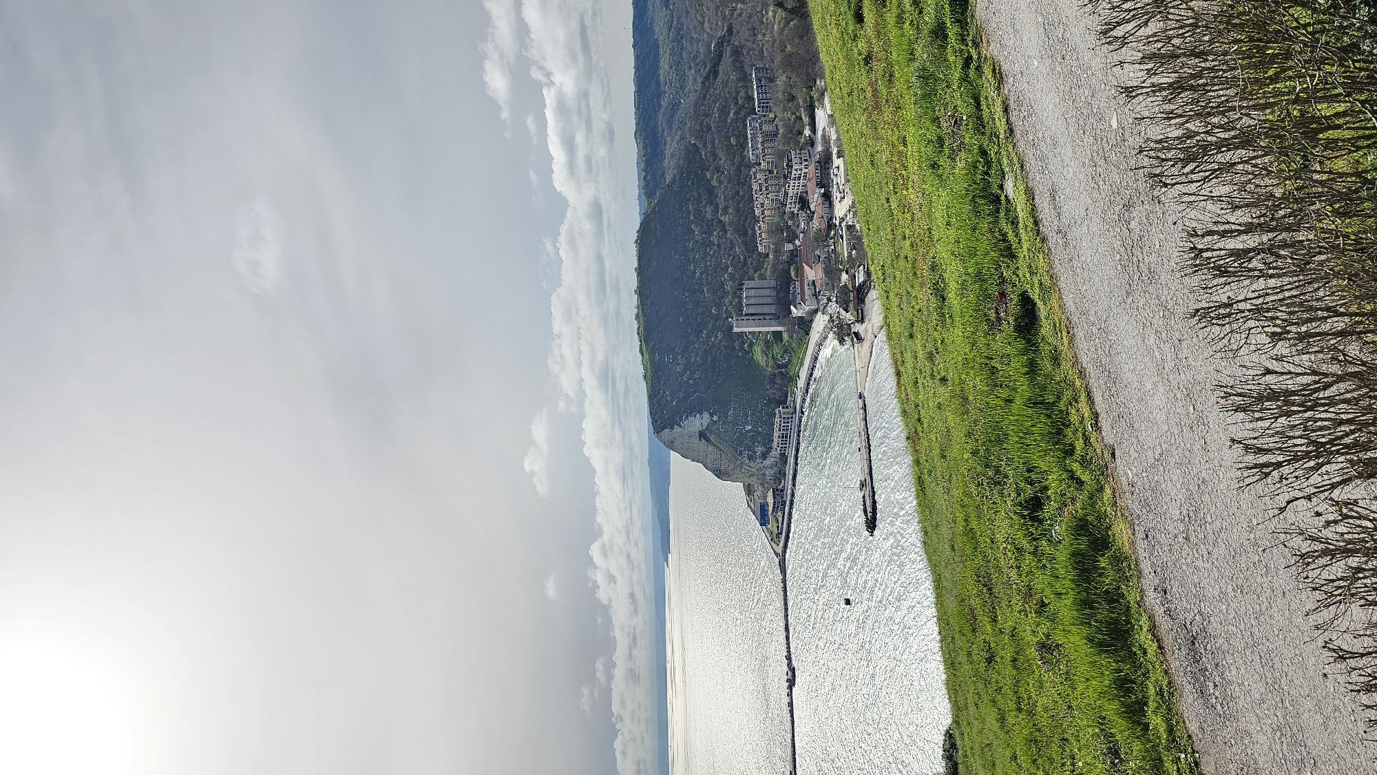 The image shows a coastal scene in Kavarna, Bulgaria, observed from an elevated position. To the right, a gray, gravel path runs along a grassy embankment. Bare branches are visible to the far right. The embankment slopes towards a beach and the Black Sea. A pier extends into the water, with buildings and a town along the coastline backed by a tree-covered hillside. The sky is overcast with white clouds. The sun appears to be partially obscured in the lower left. The water is light blue with shimmering reflections.