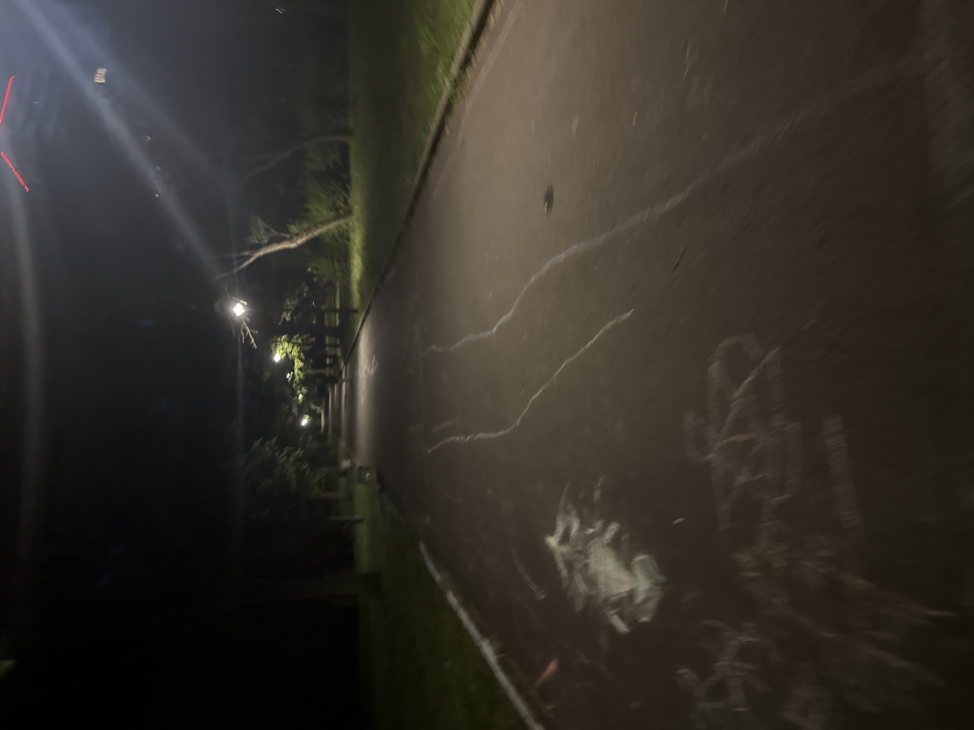 A nighttime view of a paved path in a park, likely in Burgas, Bulgaria. The path is illuminated by streetlights, casting pools of light and shadows. There are some chalk markings on the path, appearing as white and light blue lines and smudges. Trees and grass border the path, with the foliage dark due to the low light. In the distance, more streetlights and trees are visible, suggesting a well-maintained park area. The overall scene conveys a quiet, possibly deserted, park setting at night.