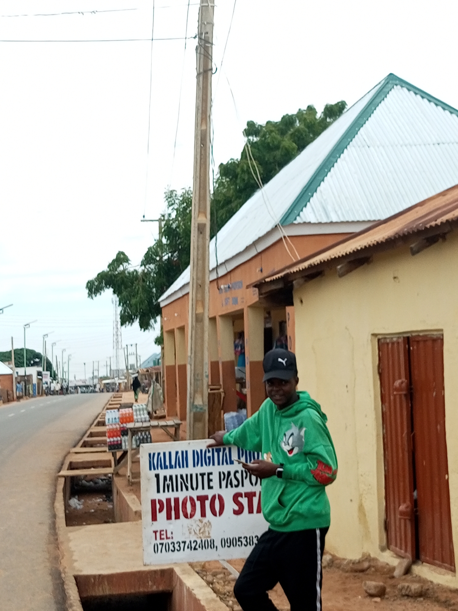 In Gombe, Nigeria, a young man wearing a green hoodie with a Tom and Jerry graphic and black pants with a white stripe stands to the right of a white sign that reads "KALLAH DIGITAL PHOTO" in black capital letters, "1 MINUTE PASPO" in smaller black capital letters, and "PHOTO ST" in large red capital letters. Below this, the sign displays "TEL: 07033742408, 0905383" in black text. The man holds a smartphone in his right hand and looks towards the camera with a slight smile. He wears a black baseball cap. The sign is positioned on a street with a concrete drainage channel in the foreground, filled with some debris. To the left of the sign, several rows of bottled drinks are stacked on shelves. In the background, a street lined with shops and trees stretches into the distance. A lamppost is visible on the left side of the street, and a tall communication tower can be seen further down. The buildings have orange and yellow walls with corrugated metal roofs. The sky is overcast.
