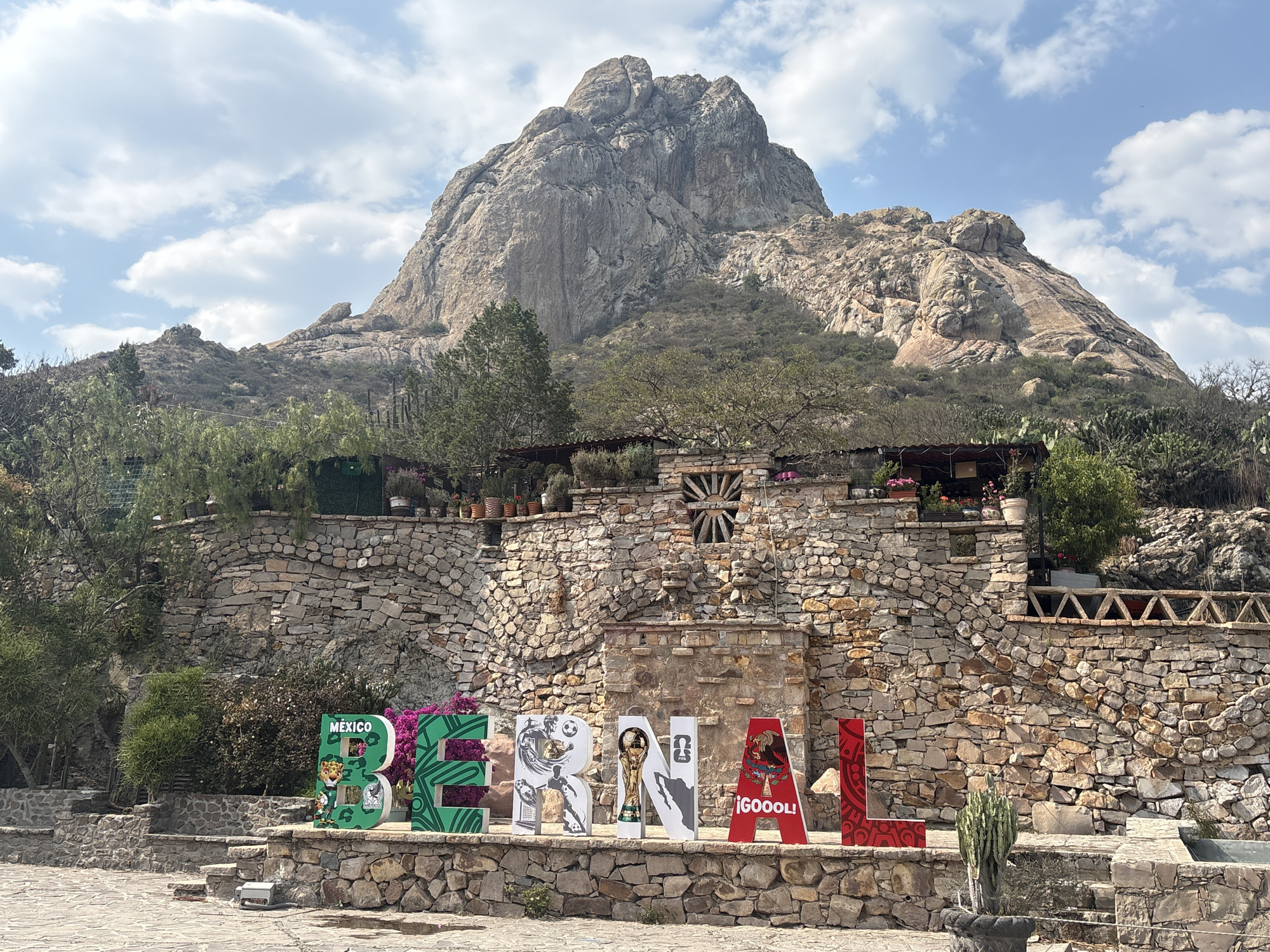The image depicts a clear day scene in Bernal, Mexico, showcasing the iconic Peña de Bernal monolith in the background. In the foreground is a stone structure with steps leading up to a platform where large, colorful letters spell out "BERNAL". The letters are decorated with soccer-themed graphics, including players, trophies, and the word "¡GOOOOL!" on one of the letters.
The stone structure features a decorative stone window and various plants. The monolith towers over the scene, its rocky surface textured by natural erosion. The sky is blue with scattered clouds, providing a bright backdrop. The foreground shows a paved area with some vegetation and stone planters.