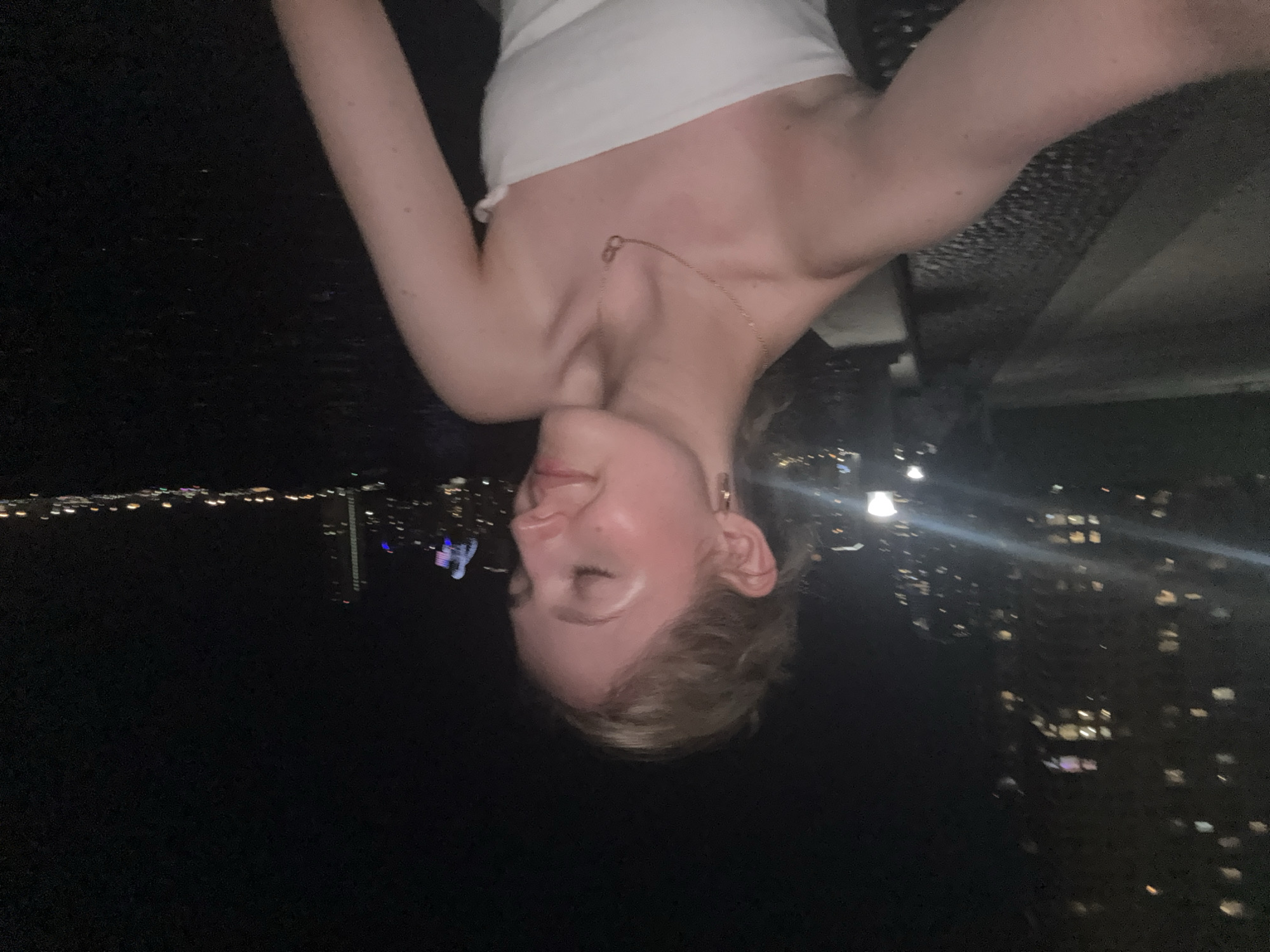 A young woman with blonde hair pulled back is captured in a selfie at night. She is wearing a white strapless top and a delicate gold necklace. Her eyes are closed, and she appears to be smiling slightly. The background shows a city skyline with illuminated buildings and lights reflecting on dark water. The scene suggests a waterfront location in Miami, likely with a view of the city across Biscayne Bay. The overall atmosphere is relaxed and serene.