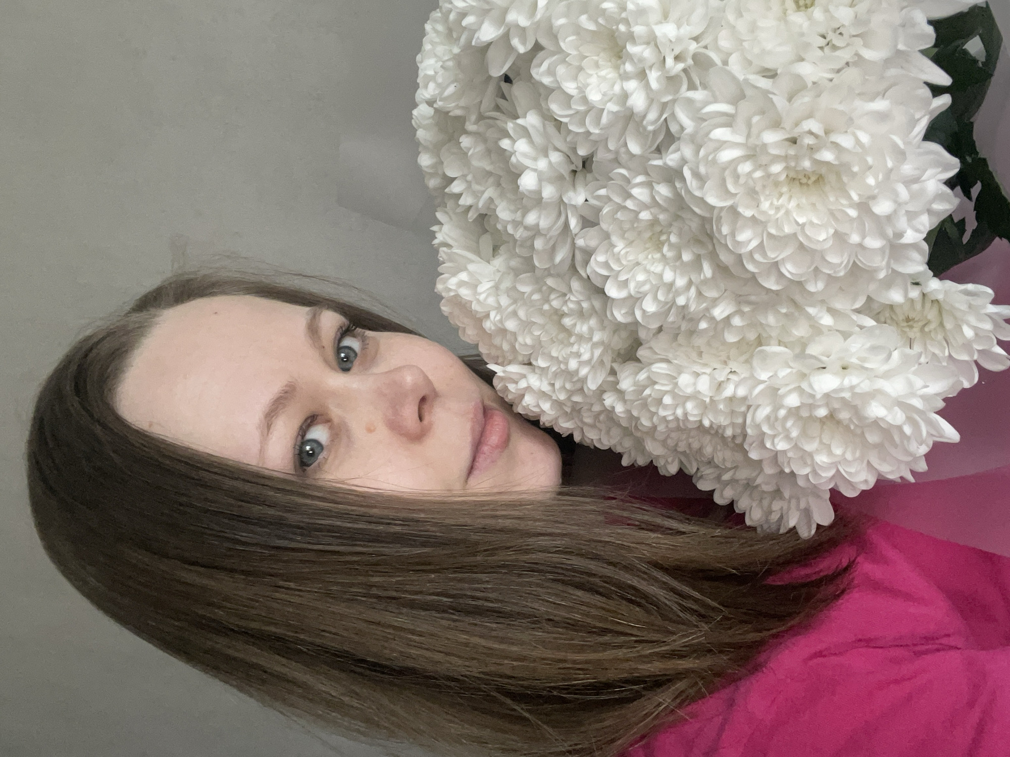 A woman with fair skin, light brown hair, and blue eyes is holding a large bouquet of white chrysanthemums. The bouquet is the focal point of the image, filling the upper right quadrant. She is wearing a bright pink top. The background is a plain, light grey wall. The angle of the photo suggests it was taken in selfie mode. The location is unknown and cannot be determined from the image.