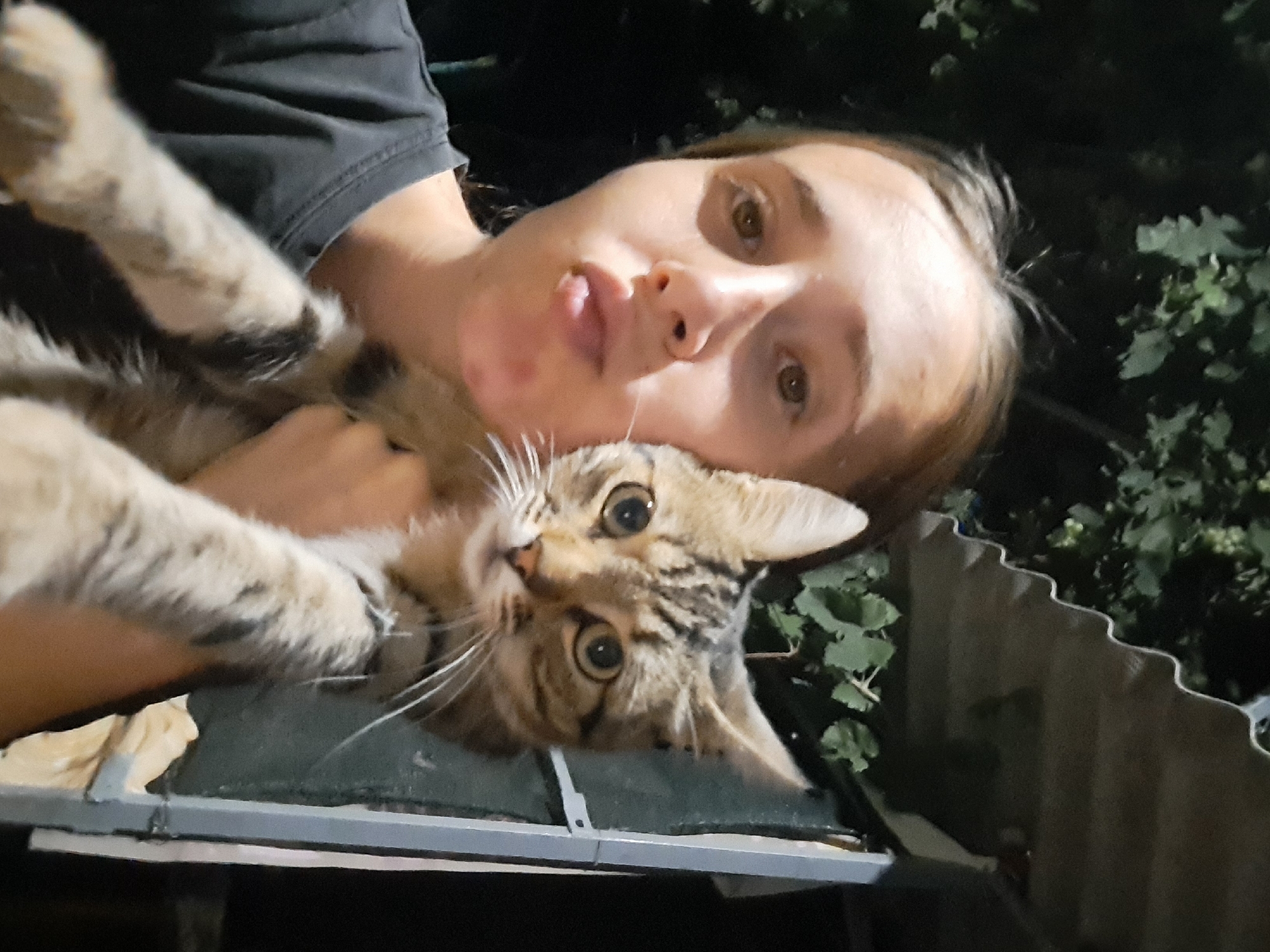 A young woman with brown hair pulled back, wearing a dark t-shirt, takes a selfie outdoors at night, holding a tabby cat. The woman is puckering her lips. The cat, with large green eyes and striped fur, is held against her face. Behind them, a dark green leafy vine climbs a structure with a corrugated roof. The background is dark, suggesting an outdoor, possibly rural or suburban, setting. The image appears to be taken in the city of Vypasne, Ukraine.