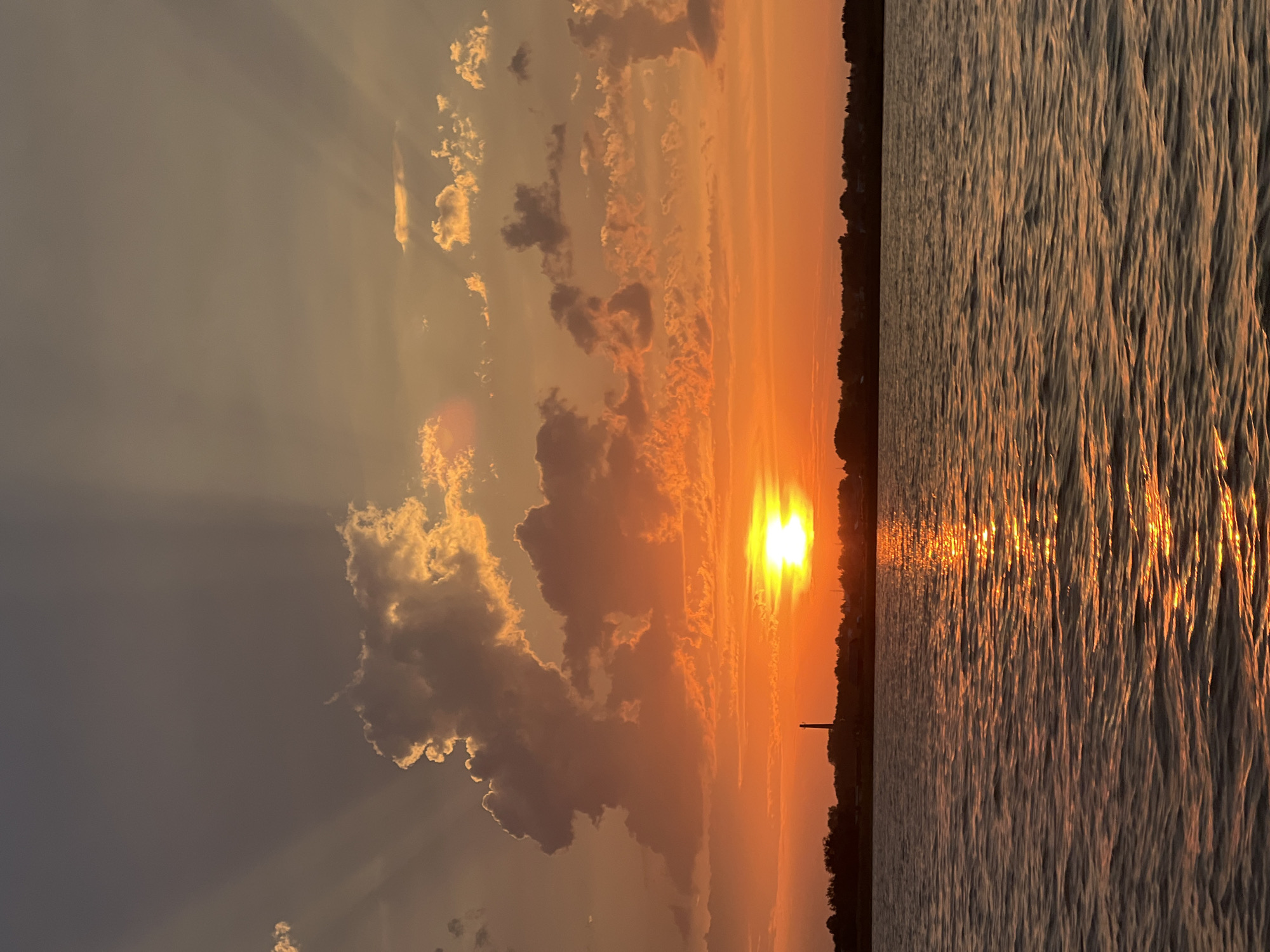 The image captures a sunset over a body of water, likely a river or lake, in the city of Novopichugovo, Russia. The sky is ablaze with warm hues of orange and yellow, with the sun appearing as a bright, radiant orb just above the horizon. Rays of light emanate from the sun, piercing through scattered clouds and creating a dramatic effect. The water reflects the vibrant colors of the sunset, with shimmering golden light dancing on its surface. In the distance, a silhouetted treeline forms the backdrop, with a slender, dark structure, possibly a chimney or tower, standing out against the colorful sky. The overall scene is tranquil and picturesque, evoking a sense of peace and natural beauty.