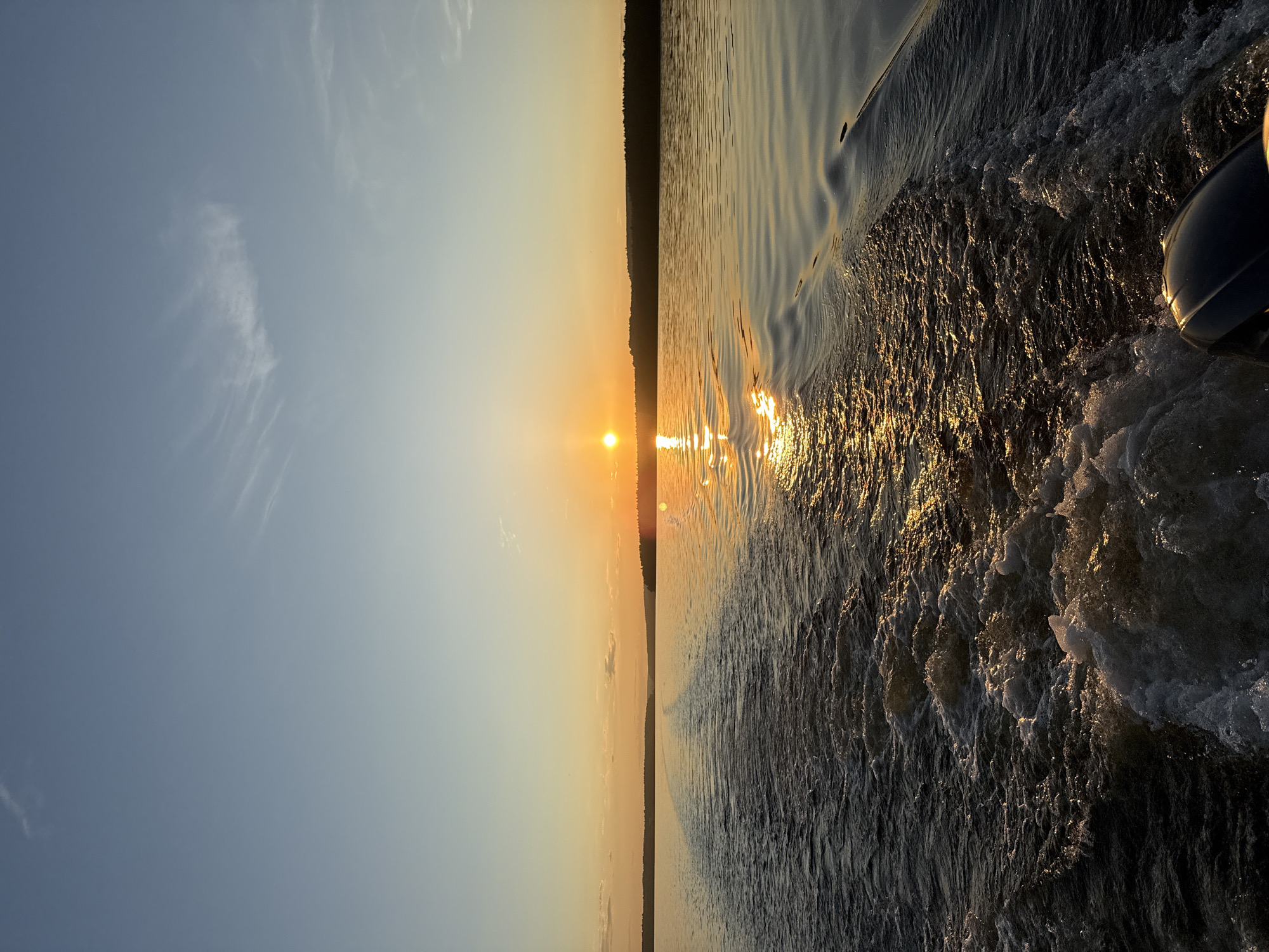 The image depicts a sunset over the Votkinsk Reservoir in Votkinsk, Russia, viewed from the rear of a moving boat. The sky transitions from light blue at the zenith to a vibrant golden-orange at the horizon, where the sun is visible as an intense orange sphere, slightly right of center. A dark, forested landmass forms a silhouette across the entire horizon. The sun's light reflects strongly on the water, creating a shimmering golden path extending from the horizon towards the lower right. The boat's wake generates turbulent white foam and waves in the foreground, forming a diminishing V-shape that recedes into the distance. The upper portion of a dark outboard motor is visible in the lower right corner, indicating the perspective. No people are explicitly visible in the scene.