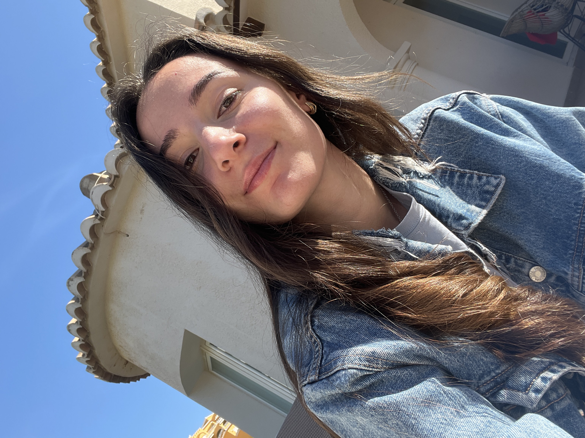 A selfie of a young woman with long brown hair, taken outdoors on a sunny day in El Poble Nou de Benitatxell/Benitachell, Spain. She is wearing a denim jacket over a light-colored shirt and is looking directly at the camera with a slight smile. The background includes a building with a white exterior and ornate roof details, along with a clear blue sky. Part of another building is visible in the lower left corner.