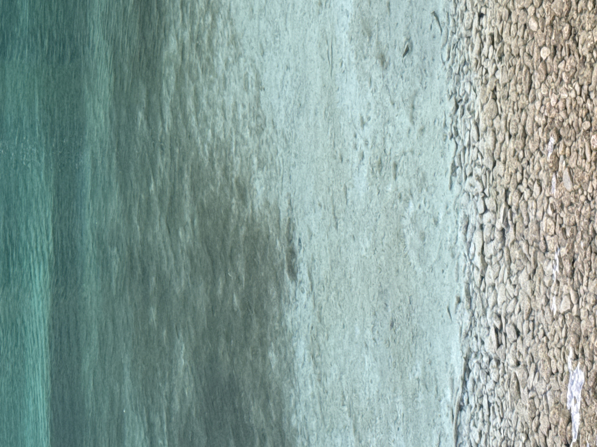 The image depicts a body of clear, shallow water with a rocky bottom, characteristic of the shoreline of the Königssee in Schönau am Königssee, Germany. The water transitions from a deeper, darker turquoise hue at the top of the frame to a lighter, almost transparent shade near the shore. The bottom is covered with an assortment of smooth, light-colored pebbles and rocks, some of which are partially submerged. The water's surface exhibits subtle ripples and reflections, indicating gentle movement. No people, animals, or man-made structures are visible in the frame. The environment is natural and serene, focusing entirely on the interaction between light and water with the rocky substrata.