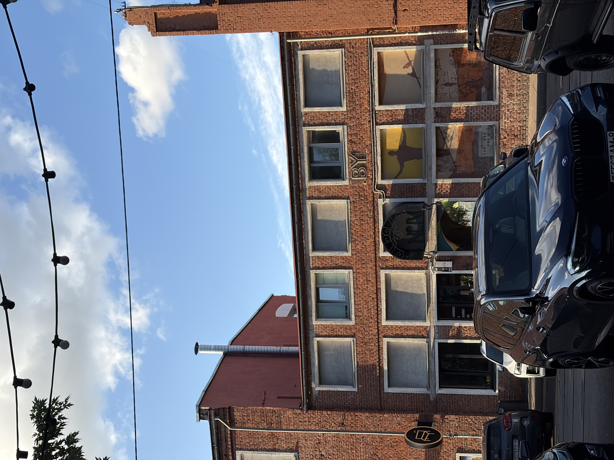 The image depicts a building facade constructed of red brick under a bright blue sky with scattered white clouds. The building has multiple windows, some covered with blinds. There is a large red structure to the left and a tall, narrow brick structure on the right. Decorative elements, including a neon sign that reads "BY" and artistic murals in some of the windows, are visible on the facade.
In the foreground, several cars are parked on a street. The most prominent is a dark blue BMW SUV in the center, facing left. To its left is a gray SUV, and to the right is a black Lada Niva with a roof rack. The cars appear to be parked on or near a street, possibly in a commercial or industrial area.
The image has been identified as being taken in Minsk, Belarus. More specifically, the location appears to be near the "Ok16" creative hub, a former factory complex in Minsk that has been repurposed into a space for art galleries, workshops, and cafes. The distinctive brick architecture, the "BY" sign, and the artistic elements on the building are characteristic of this area.