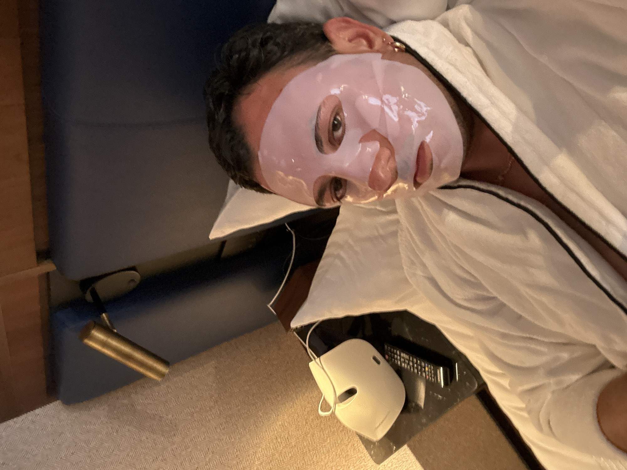 The image captures a person, likely a man with dark hair, in a close-up, reclining and wearing a translucent, pinkish sheet face mask. The mask covers his entire face, with visible cutouts for his eyes, nose, and mouth, appearing shiny and wet. He is dressed in a white terry cloth robe, partially open, revealing a gold chain around his neck. His head rests on a white pillow as he lies on a dark blue upholstered surface, possibly a bed or lounge chair, suggesting a moment of relaxation or a spa treatment. His eyes are open, looking slightly upwards.
To the right of the person's head, a dark table or nightstand is visible, holding a black remote control and a distinctive white, rounded device with a thin white cord, possibly a beauty gadget or its charger. Below this table and the reclining figure, a light brown carpet with a subtle pattern is seen. A brass-colored cylindrical fixture, possibly part of a lamp, extends from the dark blue upholstered surface on the left side of the frame. The lighting in the room is soft and warm, contributing to a relaxing indoor atmosphere. The overall scene depicts a personal self-care activity, likely in a hotel room or spa setting in Madrid, Spain. No discernible text is visible in the image.
