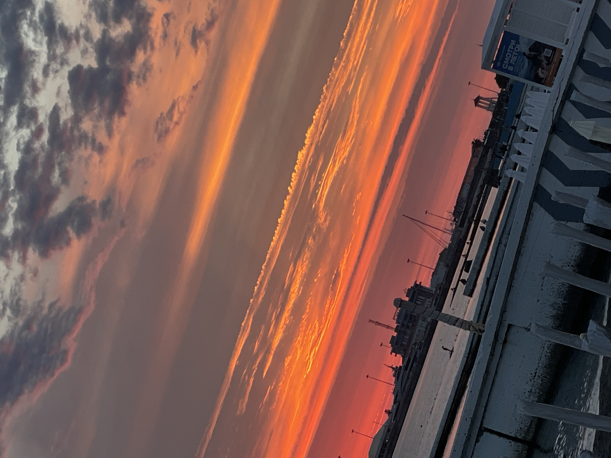 This is a medium shot of a harbor in Sochi, Russia, during a vibrant sunset. The sky is a dramatic blend of fiery orange, red, and pink streaks of clouds against a dusky gray backdrop. In the foreground, a white and blue striped railing leads the eye towards the water. Beyond the railing, the water of the harbor is calm. A white structure with a blue sign that reads "СМОТРИ В ЛЕТО" (Look into Summer) is visible on the right. In the mid-ground, several boats, including yachts and sailboats, are docked. The silhouettes of buildings and port structures are visible in the distance, along with lampposts. A person is standing on what appears to be a paddleboard in the water near the foreground. Several other figures are seated or standing along the docks in the mid-ground. The overall scene conveys a sense of leisure and natural beauty.