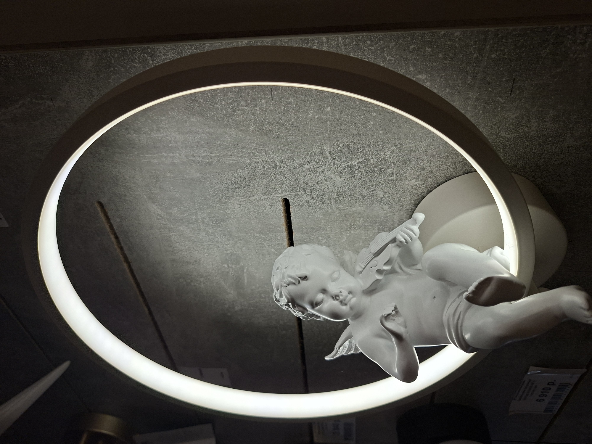 A white statue of a cherub holding a violin is placed in front of a circular light fixture against a textured gray wall. The cherub is seated, with its legs crossed and wings spread slightly. The light fixture emits a bright white glow, illuminating the cherub and casting shadows on the wall. A price tag is visible in the lower left corner of the image, showing "6 910 p." The background appears to be an interior setting, possibly a store or display area. The image was taken in Izhevsk, Russia.