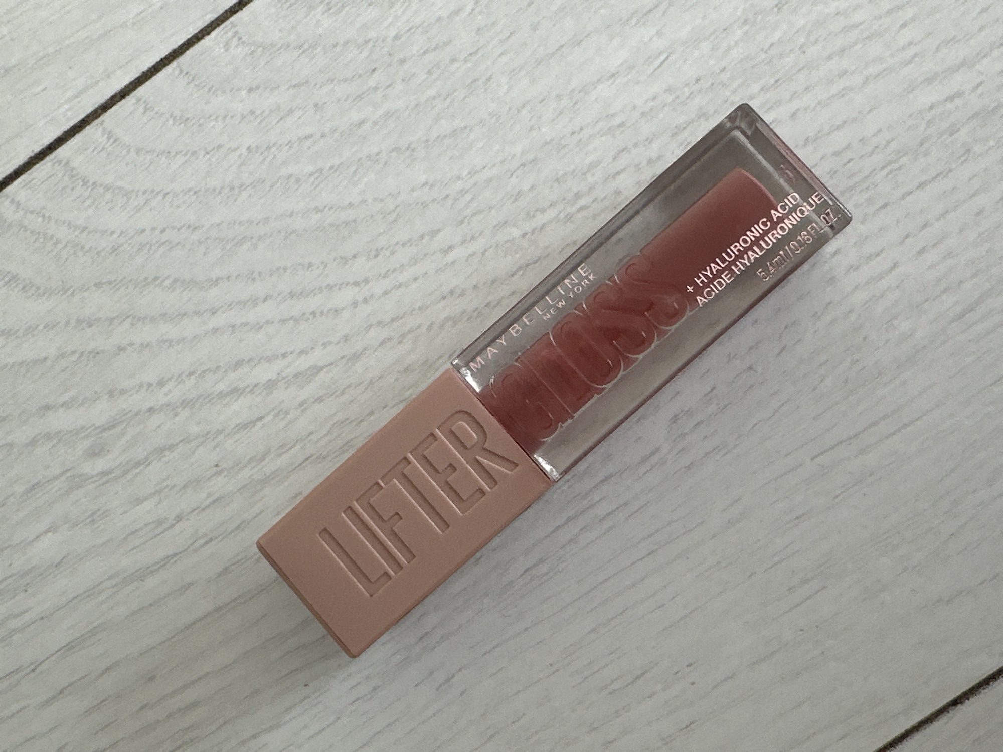 A Maybelline Lifter Gloss is resting on a light wood-grain surface. The gloss is in a clear tube with a pink cap that says "LIFTER" in embossed letters. Inside the tube, the gloss is a peachy-pink color. Text on the tube reads "MAYBELLINE NEW YORK" and "+ HYALURONIC ACID ACIDE HYALURONIQUE 5.4ml / 0.18 FL.OZ." The background is a light-colored, wood-grain surface with visible lines and grain.