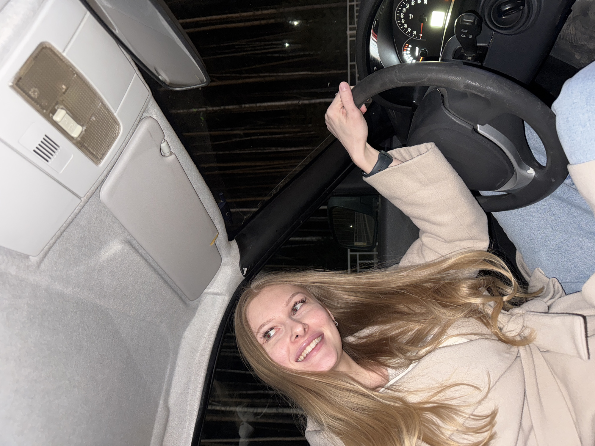 A young blonde woman is captured inside a car, smiling widely and looking upwards to her left. Her long hair flows around her, suggesting movement or a breeze. She wears a light-colored coat, and her right hand, adorned with a black smartwatch, rests on the steering wheel. Her expression is cheerful and happy.
The interior of the car is visible, featuring a light-colored headliner and an overhead console with an interior light and a vent. A sun visor is partially visible. The dashboard features a speedometer displaying readings such as 0, 20, 40, 60, 80, 100, 120, 140, 160, 180, and 200, likely indicating km/h, with the needle resting at zero. A gear shift lever is also visible. The steering wheel appears well-used. Through the car's windows, a dark background with distinct horizontal lines suggests a nighttime setting, possibly a multi-story parking garage or an urban environment at night. The car appears to be stationary. This scene takes place in Perm, Russia.