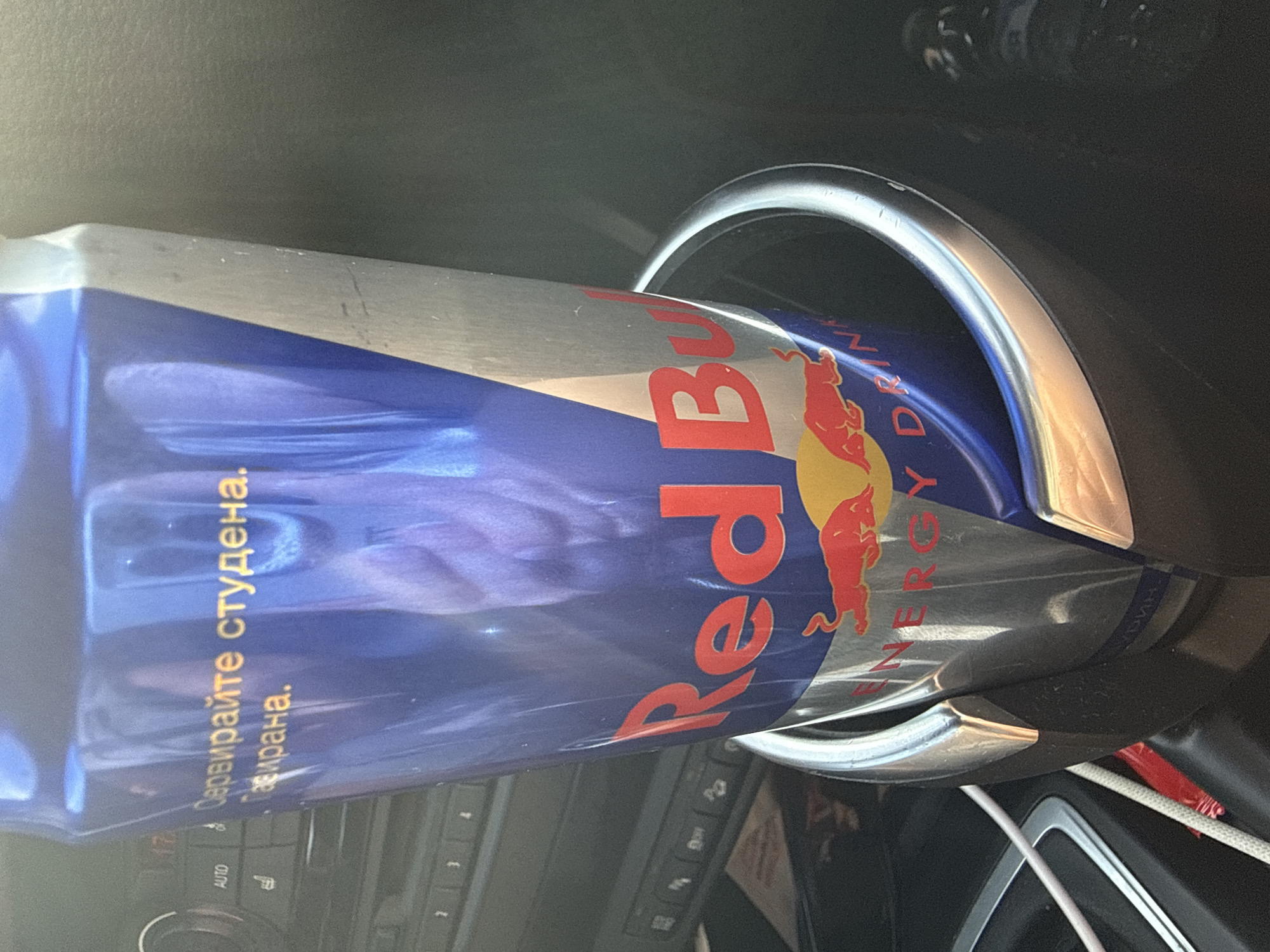A Red Bull energy drink can is held in a car's cup holder. The can is blue and silver with the Red Bull logo. Text in Cyrillic script is visible on the can. The car's interior is visible in the background, including buttons and a screen. The scene appears to be in Polsko Padarevo, Bulgaria.