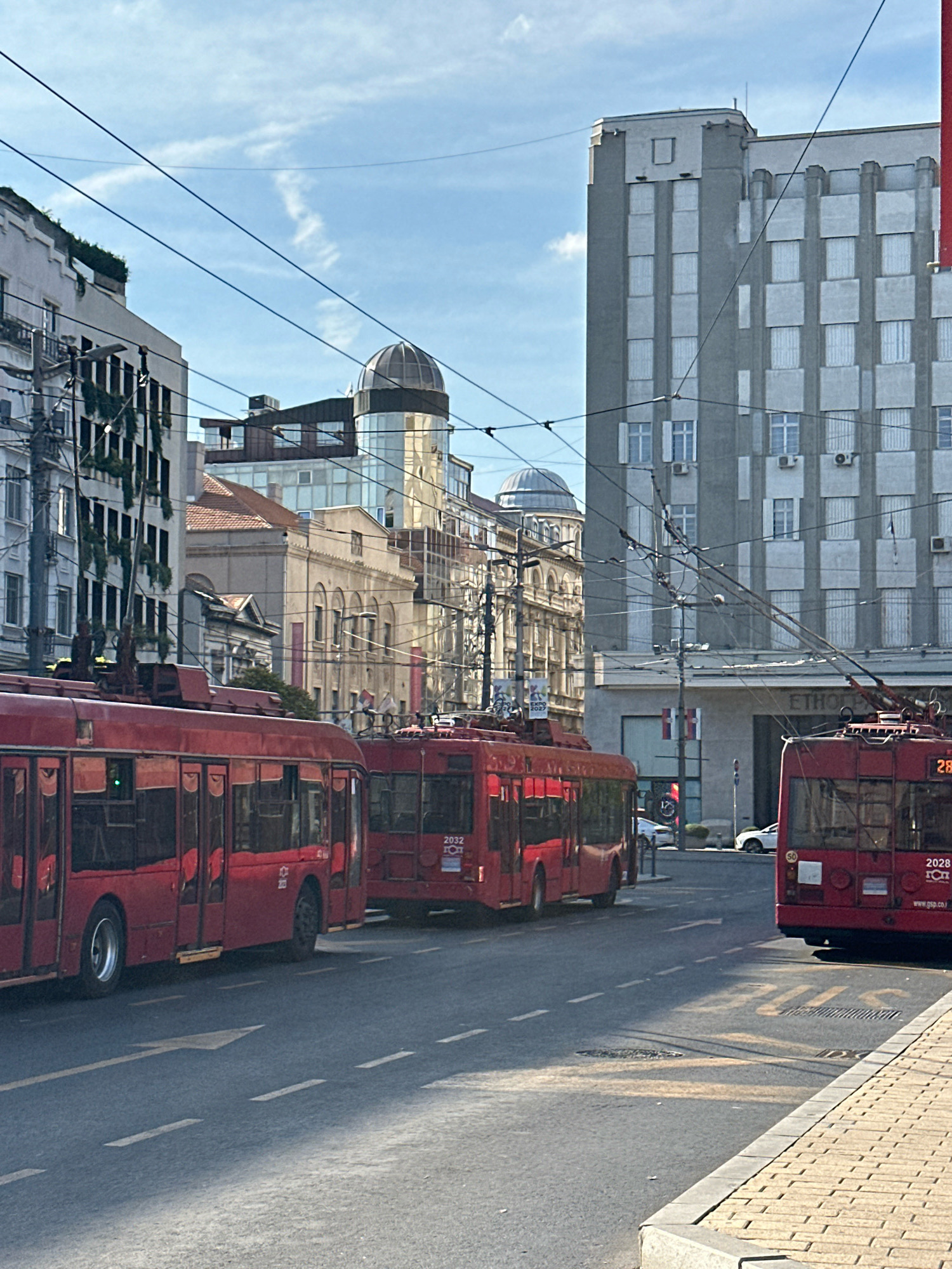 The image shows a street scene in Belgrade, Serbia, likely captured on a sunny day. In the foreground, three red trolleybuses are parked or moving slowly on a paved road marked with white lane lines and a faded yellow "BUS" marking. The trolleybuses have reflective windows and are powered by overhead wires, indicated by the poles extending from their roofs.
Behind the trolleybuses, a series of buildings line the street. The buildings display a mix of architectural styles, including a modern grey building with a grid of windows on the right, and older buildings with ornate facades and domes in the background. Some of the buildings have visible air conditioning units. Overhead, a network of electrical wires crisscrosses the sky.
The environment appears to be a commercial or urban district, with sidewalks and streetlights visible. The overall impression is of a bustling city street during daytime, with public transportation playing a prominent role. No people are clearly visible in the image.