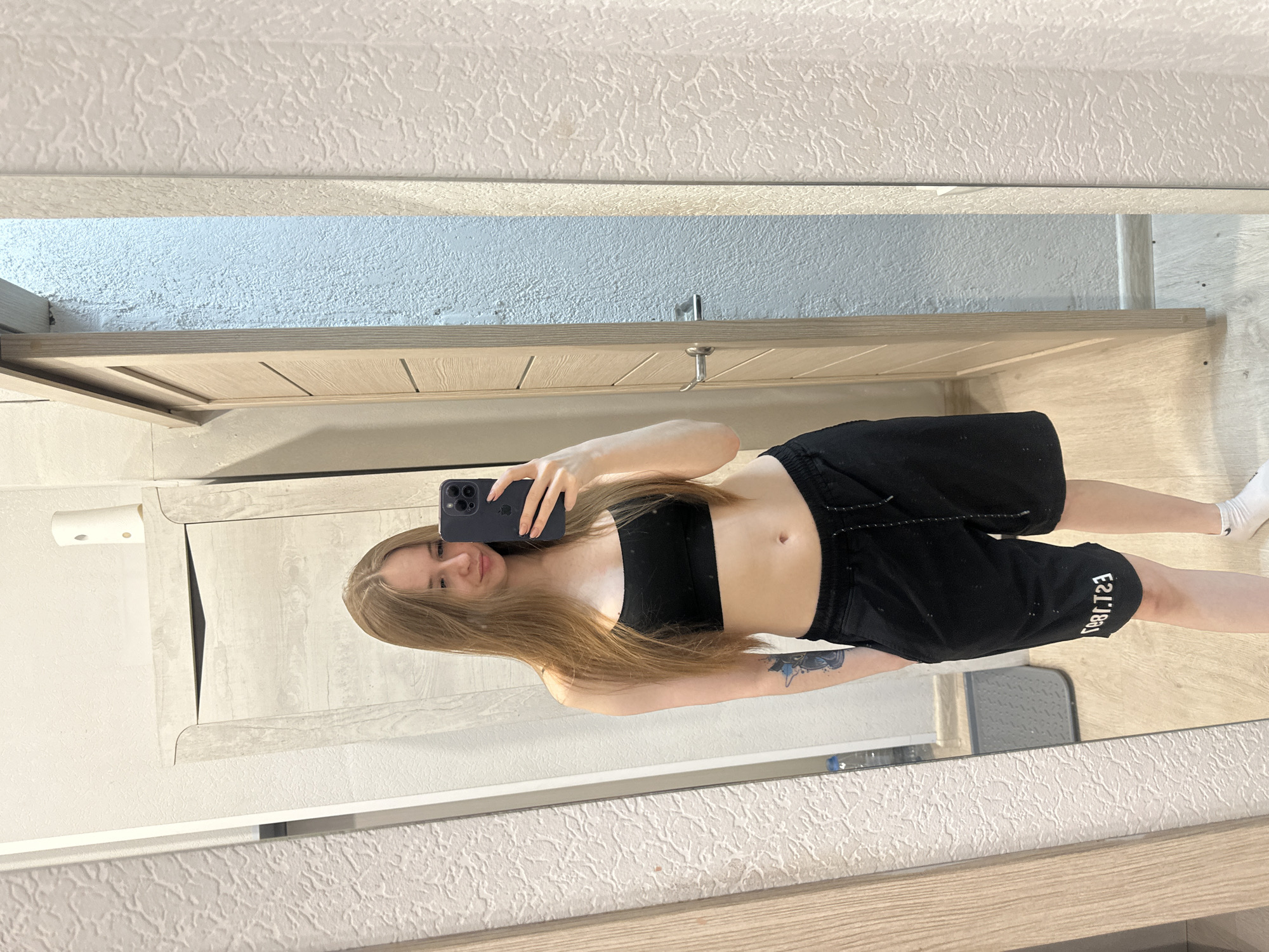 A full-length image shows a young woman in a mirror selfie, against a backdrop of a bedroom or dressing area. She is positioned in the center of the frame, holding a smartphone in her right hand to capture the reflection.
She is wearing a black sports bra and matching black athletic shorts. Her long blonde hair falls down her chest. Her skin appears pale, and a tattoo is visible on her left forearm. She is wearing white socks.
The reflection in the mirror indicates a neutral colored wall and furniture with light wood-grain finish. In the background, a door or cabinet is visible, adding depth to the setting. The floor has a light-colored wooden texture. A small grey step stool is visible.
The image appears to be taken indoors in a private residence, but further details about the location in the city of Unknown, Unknown are not possible to derive.