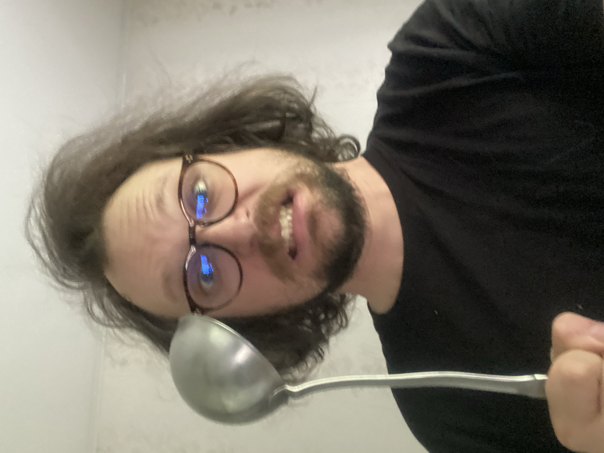 A bearded man with glasses holds a metal ladle in front of his face. He has curly brown hair and is wearing a black t-shirt. The background appears to be a white or light-colored wall. There is no indication of Rusanovo, Russia.