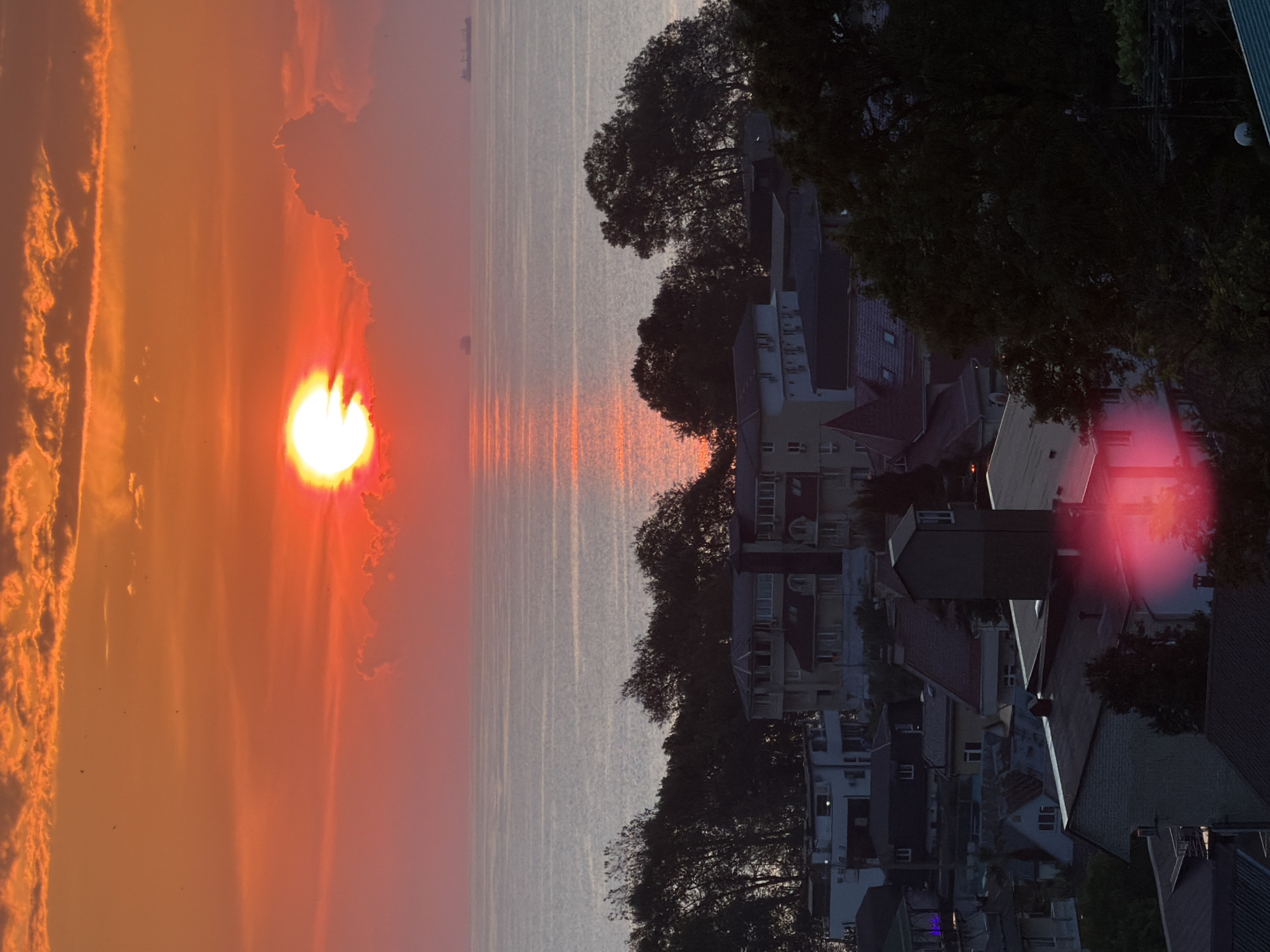 The image depicts a vibrant sunset over the Black Sea, viewed from a hillside overlooking the coastal city of Sochi, Russia. The sun, a bright orb of fiery orange, is partially obscured by wispy clouds, casting a warm glow across the sky and the rippling water. The sea is calm, with subtle reflections of the sunset painting shimmering streaks of light. In the distance, two cargo ships are visible on the horizon.
The foreground of the image is dominated by a dense canopy of dark green trees, silhouetted against the bright sky. Nestled amongst the trees are a cluster of buildings, appearing to be residential or small hotels, with various architectural styles ranging from modern to more traditional. Rooftops are visible, some with red tiles and others with darker materials. The buildings are tightly packed together, suggesting a dense urban development along the hillside.
In the lower part of the image, there is a noticeable red lens flare, which appears to be a photographic artifact rather than a part of the natural scene. The overall impression is one of a picturesque coastal town at dusk, with the natural beauty of the sunset contrasting with the man-made structures.