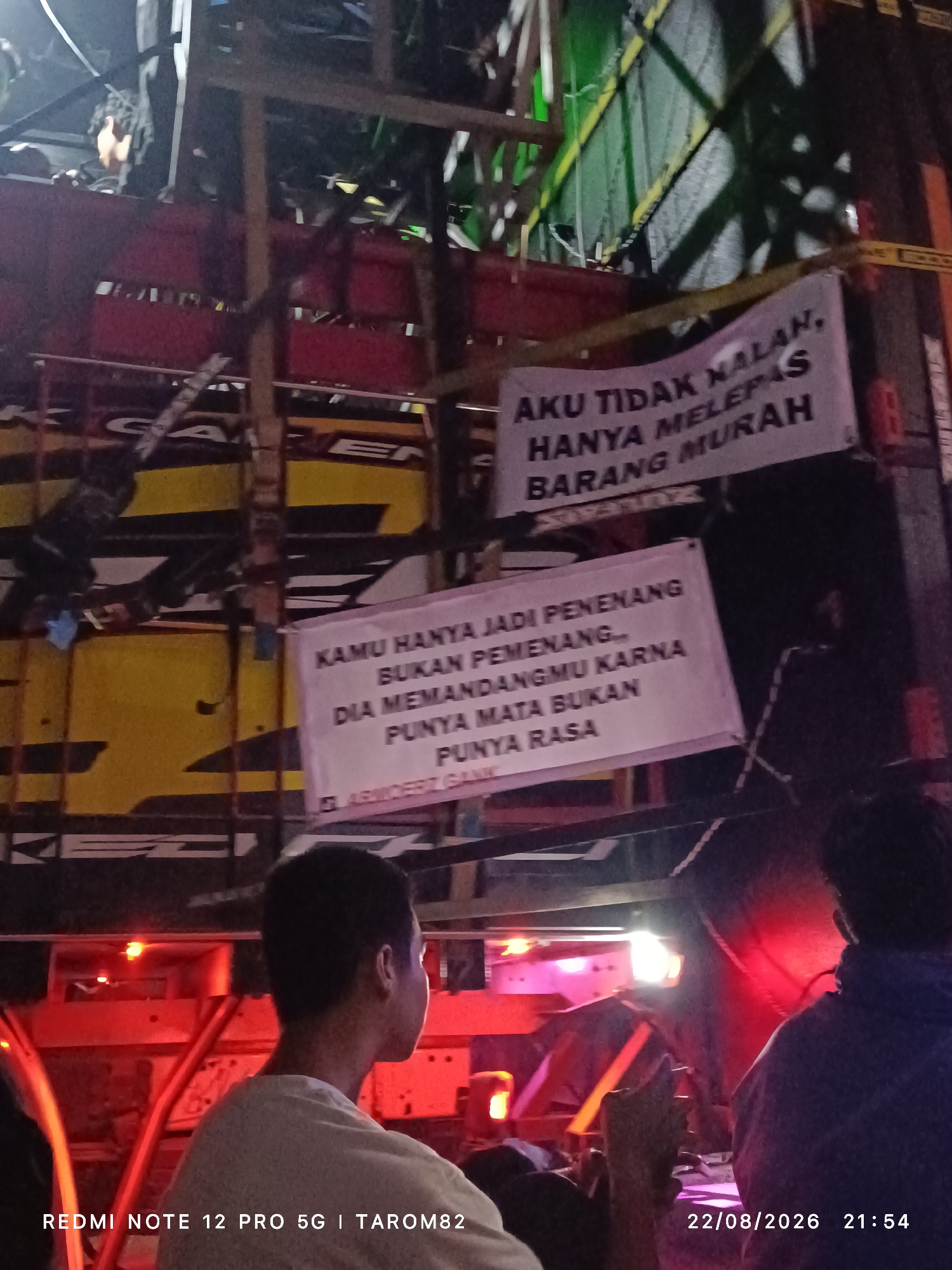 The image depicts a nighttime scene at what appears to be an outdoor event or gathering in Mundusewu, Indonesia. In the foreground, two individuals are partially visible, facing away from the camera. One person, on the left, is wearing a light-colored t-shirt and looking towards a truck. The other person, on the right, is wearing a dark hooded jacket.
Dominating the background is a large truck adorned with banners. One banner, hanging from the top of the truck, displays text in Indonesian: "AKU TIDAK NALAN, HANYA MELEPAS BARANG MURAH" which translates to "I'm not crazy, I just sell cheap goods." Below this, another banner reads: "KAMU HANYA JADI PENENANG BUKAN PEMENANG DIA MEMANDANGMU KARNA PUNYA MATA BUKAN PUNYA RASA" which translates to "You are just a winner, not a champion. He looks at you because he has eyes, not because he has feelings."
The truck itself has yellow and red detailing. The overall environment is dimly lit, with the primary light sources appearing to be the lights from the truck and possibly some ambient lighting from the event. The image is captured with a date and time stamp in the bottom right corner: "22/08/2026 21:54". In the bottom left corner, there is text: "REDMI NOTE 12 PRO 5G | TAROM82".