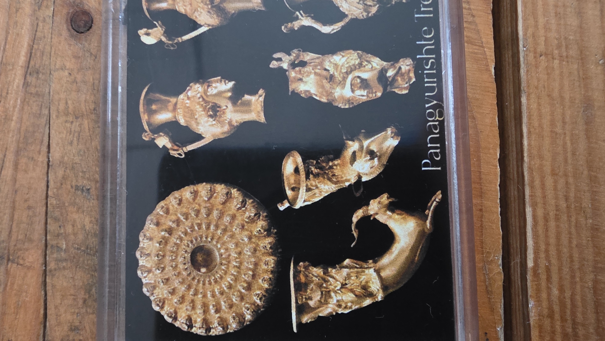 The image captures a close-up view of a photograph displaying ancient golden artifacts, resting on a textured wooden surface. The photograph is visible through a clear, reflective protective layer, possibly glass or plastic.
The central subjects in the photograph are several ornate golden objects set against a dark, likely black, background. These include multiple golden drinking vessels, known as rhyta, which appear to be sculpted in the form of animal heads or mythical creatures, exhibiting intricate details. One prominent object is a large, circular golden plate or phiale, characterized by its concentric rings of raised, intricate patterns.
Vertically oriented text is partially visible on the right side of the photograph, clearly reading "Panagyurishte Tre". This indicates that the artifacts depicted are part of the renowned Panagyurishte Treasure, a significant archaeological find from Thracian antiquity.
No specific activity or event is taking place, beyond the presentation of these historical artifacts within the photograph. The wooden surface, with its visible grain and warm brown tones, frames the photograph. Given the text on the photograph, the artifacts originate from Bulgaria, aligning with the provided location context of Blagoevgrad, a city in Bulgaria. The lighting appears to be indoor and artificial, illuminating the golden luster of the artifacts.