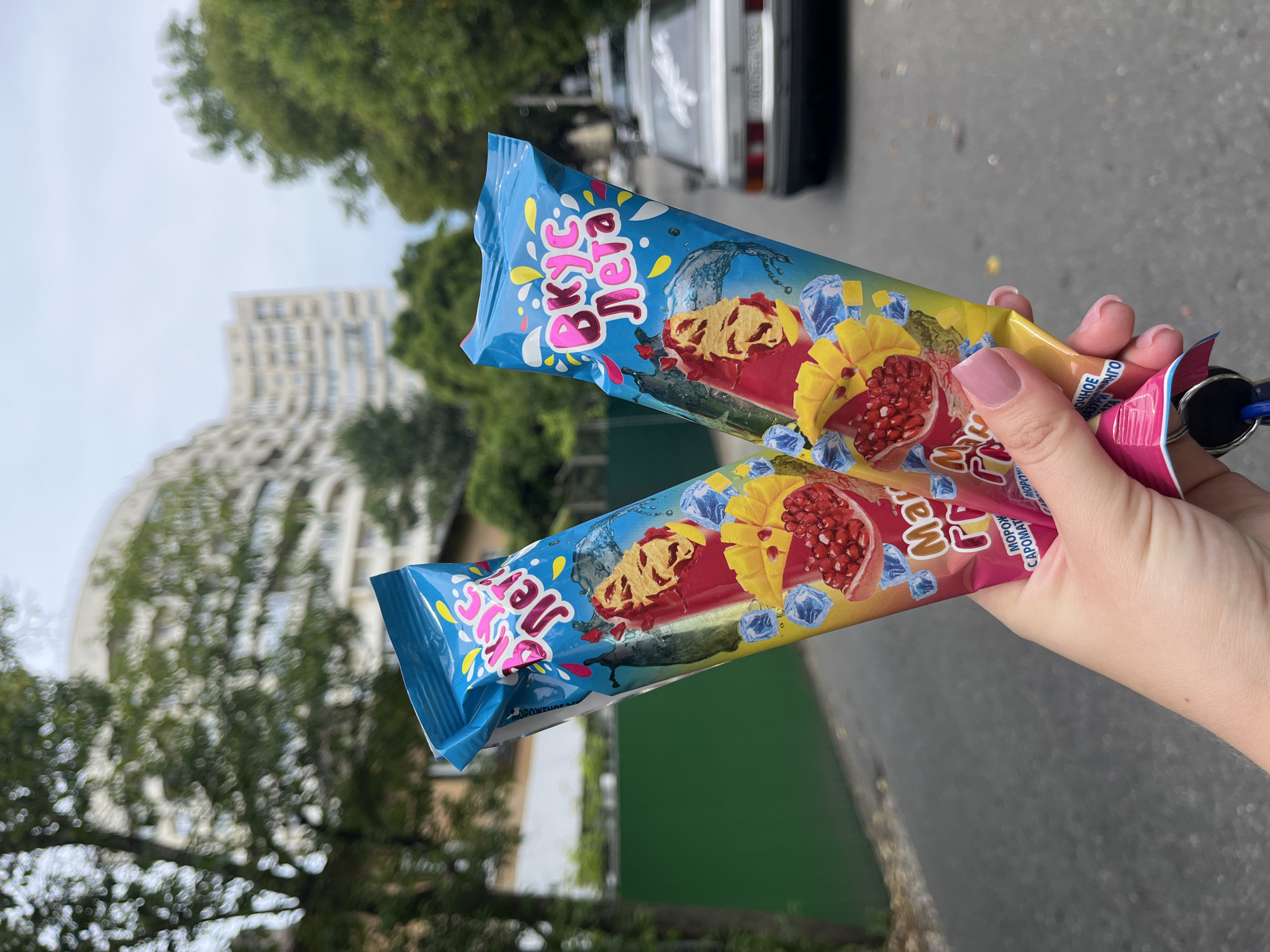 A person's hand holds two popsicle wrappers with blue, pink, and yellow designs depicting ice cubes, water splashes, and fruit slices of pomegranate and mango. The text on the wrappers reads "Вкус Лета" (Taste of Summer) in stylized pink and yellow lettering. Below this, in smaller text, it says "Манго Гранат" (Mango Pomegranate) and "МОРОЖЕНОЕ" (Ice Cream). The background is slightly blurred, showing trees, a building, and a white car on a paved street. The scene appears to be outdoors in a city.