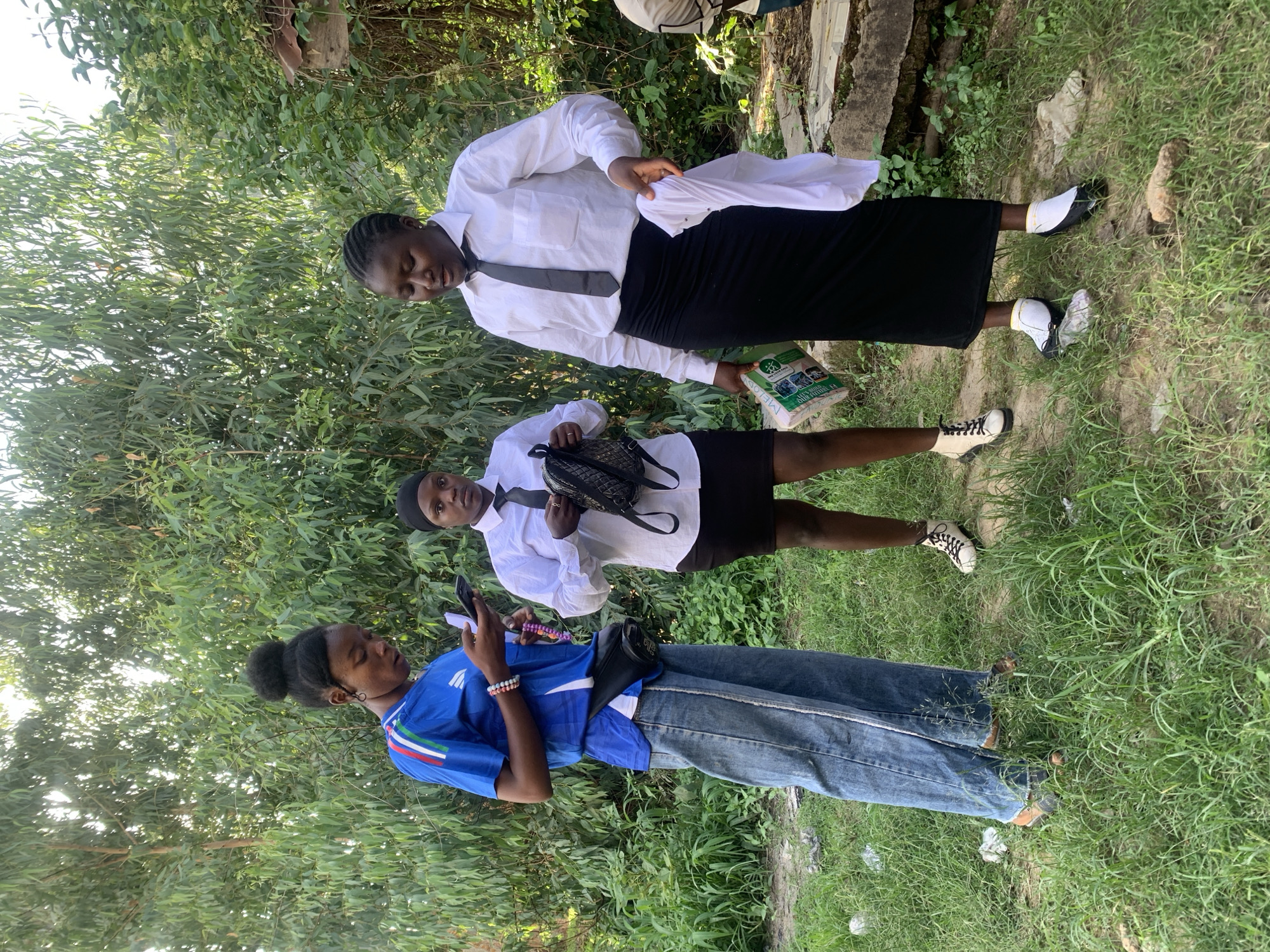 This image, captured outdoors in Jos, Nigeria, depicts three young adult females standing amidst dense greenery. The ground is covered in short green grass, interspersed with patches of bare soil and small rocks. The background features a thick canopy of trees and undergrowth, with sunlight filtering through the leaves from the upper left. A concrete block and a small, weathered wooden structure entangled with vines are visible on the far right.
From left to right:
The first individual wears a blue short-sleeved jersey with white, green, and red stripes on the shoulders, a white undershirt, and light blue wide-leg denim trousers with white vertical stripes along the outer seams. She has dark skin and her hair is styled in a high bun. A black fanny pack is worn diagonally across her chest. She wears multi-colored beaded bracelets on her right wrist and sandals on her feet, partially obscured by grass. She is holding a smartphone in both hands and appears to be actively viewing its screen, also holding a string of purple beads in her left hand.
The second individual, positioned centrally, has dark skin and wears a black head covering, a white long-sleeved collared shirt, a black tie, and a black mini-skirt. She is wearing white and black lace-up boots. She holds a black quilted backpack in her left hand and is looking slightly towards the right side of the frame.
The third individual, on the right, has dark skin and her hair is styled in cornrows. She wears a white long-sleeved collared shirt, a black tie, a black maxi skirt, and black and white flat shoes with white socks. She holds a folded white garment in her right hand and a rectangular green and white package labeled "MELS" in her left hand. She is looking downward and to her left, seemingly at the package or the garment.
All three individuals are standing in a casual arrangement, with minimal direct interaction visible between them.