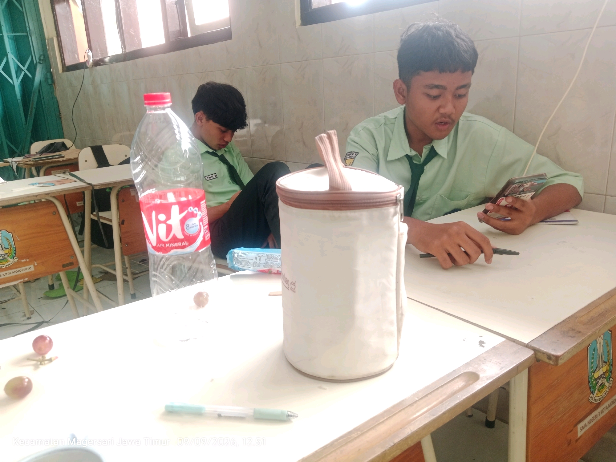 Two male students in light green school uniforms sit at desks in a classroom. The student on the right holds a smartphone and appears to be looking at it. The student on the left is looking down. A large white cylindrical container with a brown lid and handles sits on the desk in the foreground. A clear plastic bottle of Vit A. Mineral water is to the left of the container. Several grapes and a pen are also on the desk. The room has tiled walls and windows with blinds. The scene appears to be in a school in Magersari, East Java, Indonesia, as indicated by the timestamp and text overlay.