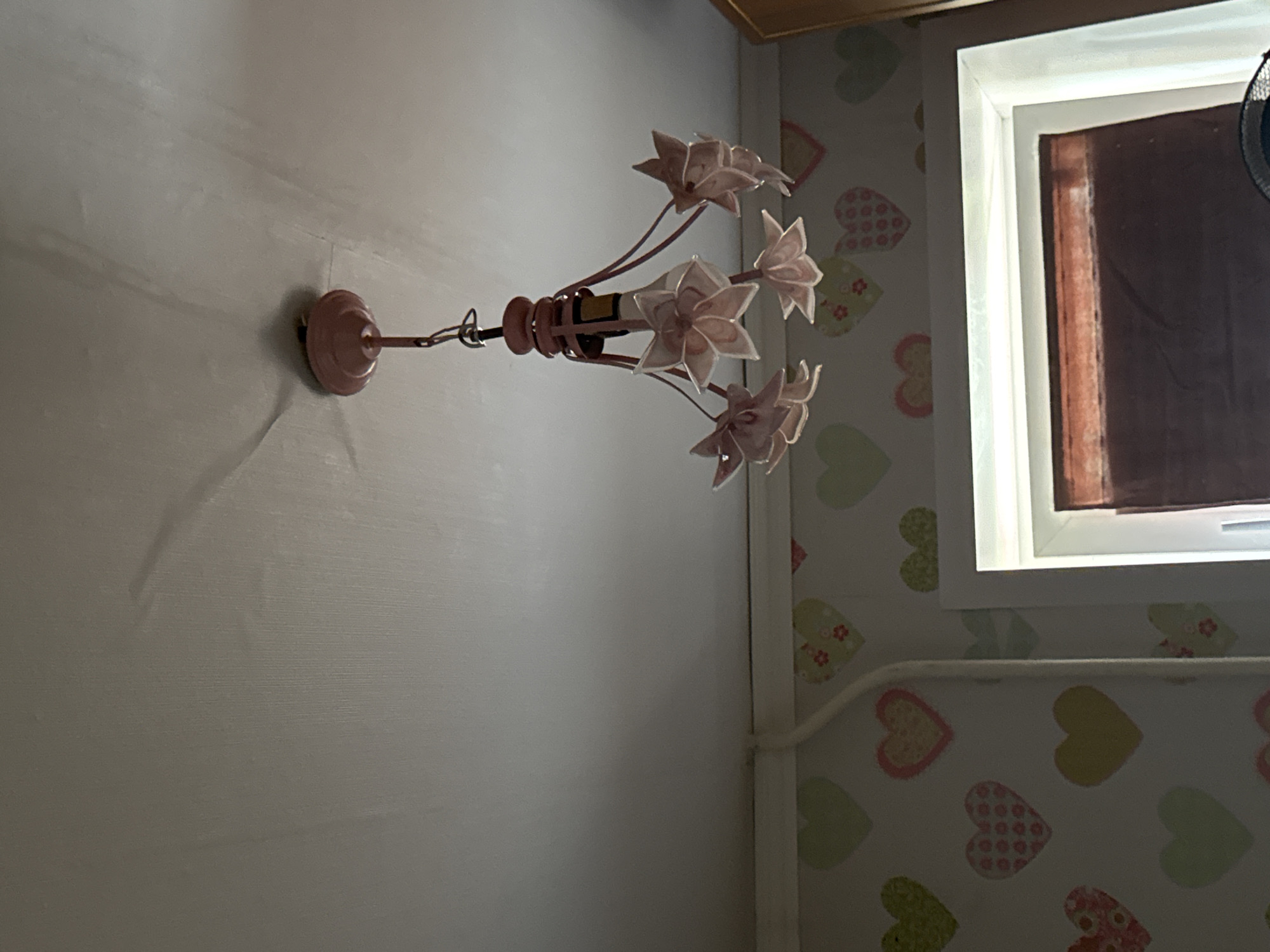 The image depicts a pink floral chandelier hanging from a white textured ceiling. The chandelier has multiple flower-shaped light fixtures made of a translucent pink material, suspended by thin wires. Below the ceiling, a wall is visible, adorned with wallpaper featuring a pattern of colorful hearts in various shades of pink, yellow, and green. A white electrical conduit runs horizontally along the top of the wallpapered wall. In the lower right portion of the image, a white window frame is visible, with a dark-colored blind partially obscuring the view outside. The scene appears to be indoors, likely in a residential setting, in the city of Yaromincki Selski Savet, Belarus. No people are present in the image.